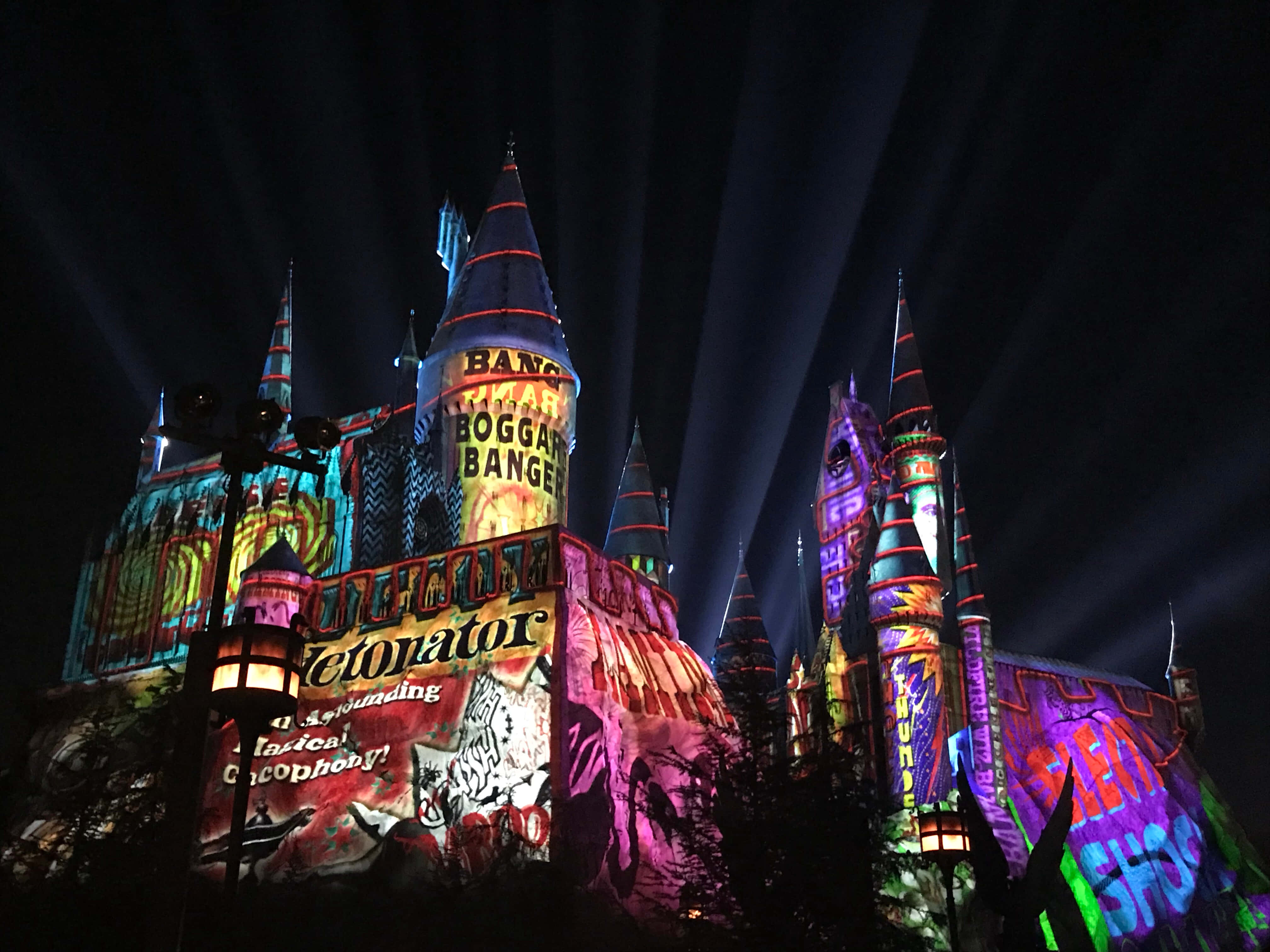Christmas At Hogwarts 桌布 壁紙 — 夜晚的城堡被充滿活力的魔法投影和戲劇性的聚光燈照亮，光束劃過黑暗的天空。