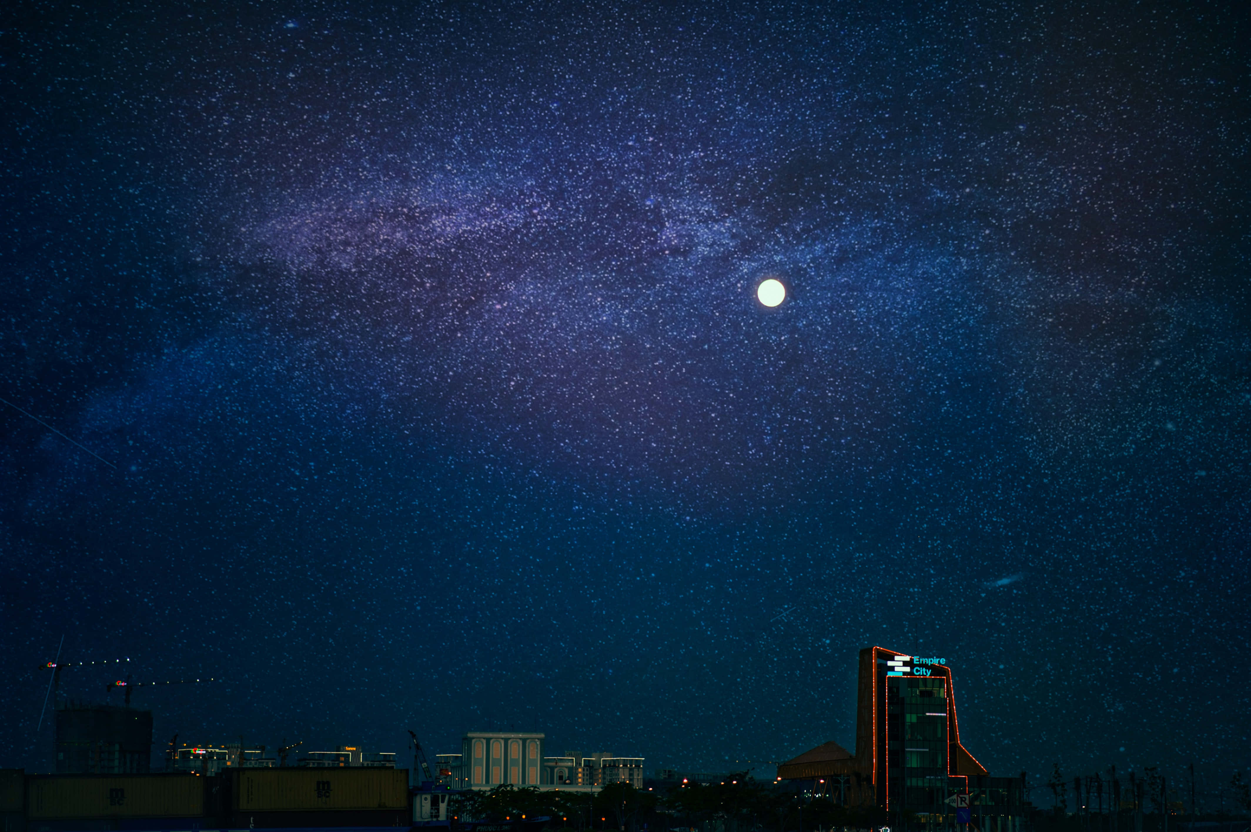 月光 桌布 壁紙 — 夜晚的城市天際線，下方是充滿活力的星空，明亮的月亮和銀河拱起。