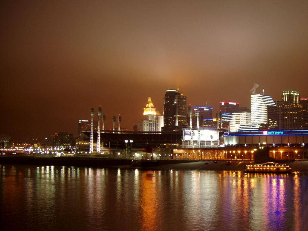 夜空 桌布 壁紙 — 夜晚的城市景觀，在朦朧的天空下，平靜的水面倒映著燈火通明的摩天大樓和發光的體育場。