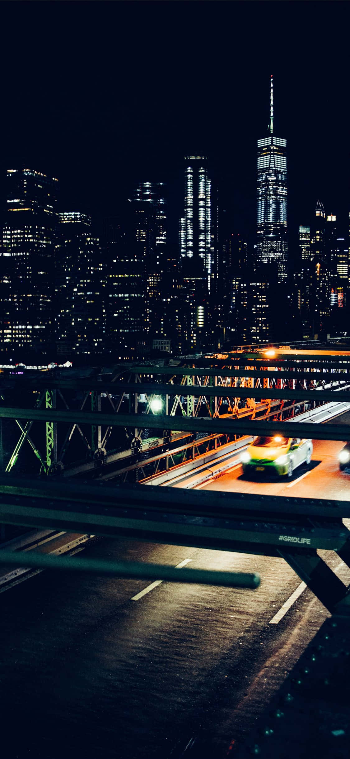 iPhone Xs New York 桌布 壁紙 — 夜晚的城市景觀，有一座亮燈的橋樑和一輛黃色計程車在摩天大樓下疾馳而過。