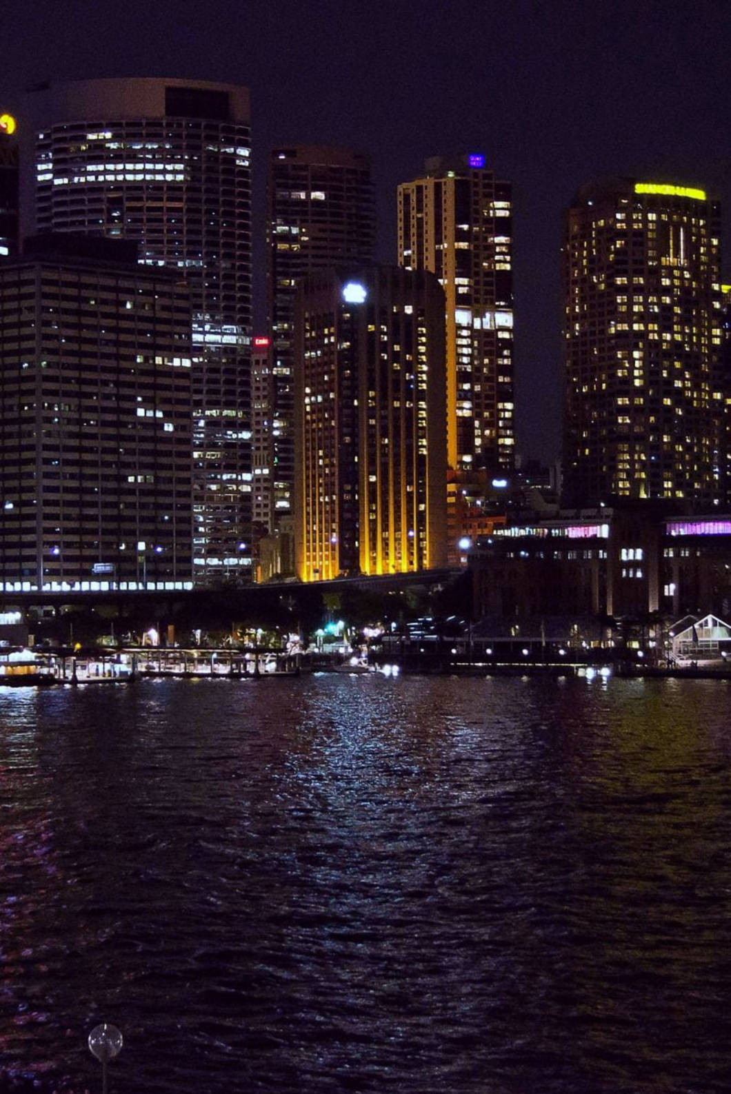 iPhone 桌布 壁紙 — 夜晚的城市景觀，燈火通明的摩天大樓在深藍色天空下的黑暗水面上倒映。