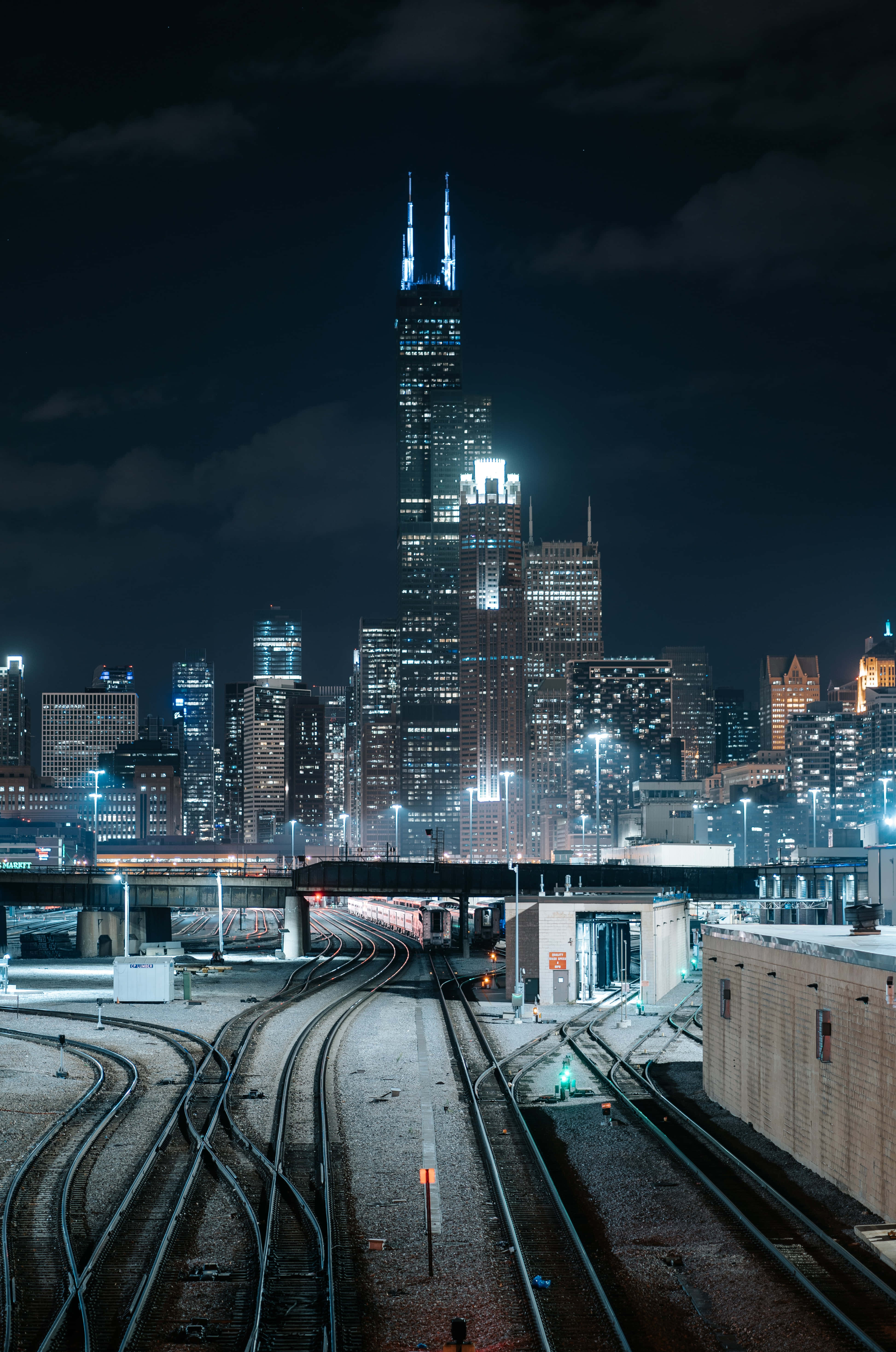 Chicago City Night 桌布 壁紙 — 夜晚的城市景觀，特色是燈火通明的摩天大樓和匯聚的火車軌道，在黑暗的天空下。