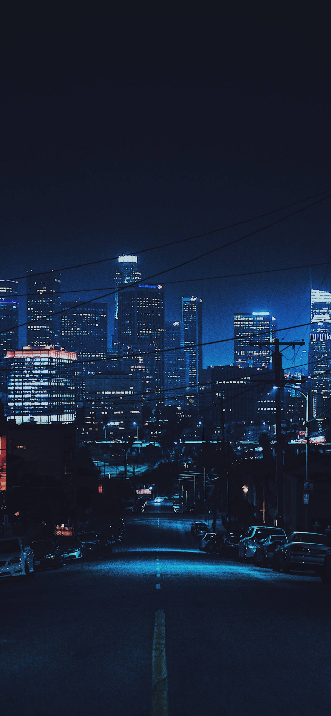 Skyscraper 桌布 壁紙 — 夜晚的城市景觀，閃爍的摩天大樓和一條黑暗的街道，兩旁停滿了汽車，在深藍色的天空下。