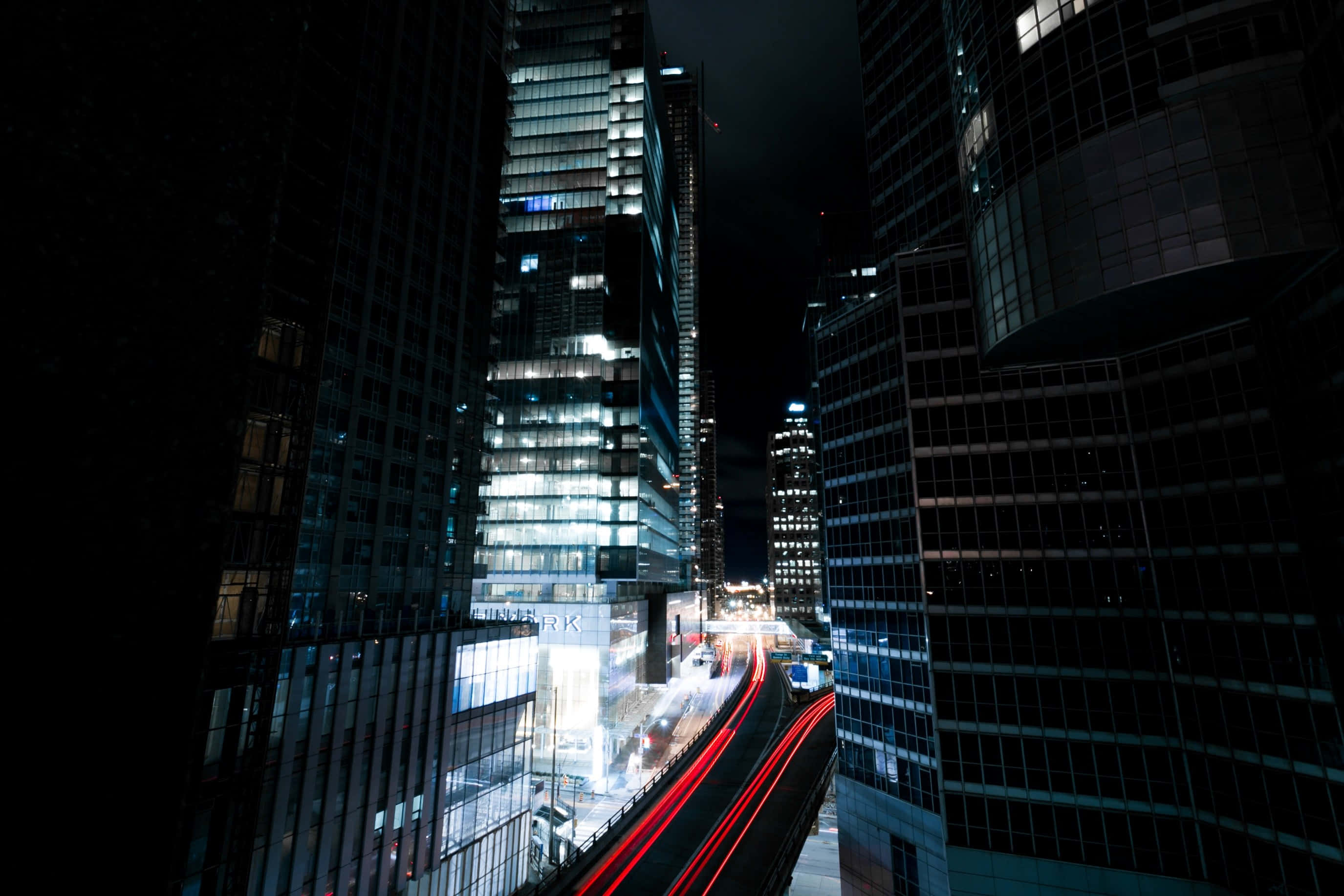 Lit iPhone 桌布 壁紙 — 夜晚的城市景觀，高聳的玻璃摩天大樓和下方移動車輛發出的紅色光跡。
