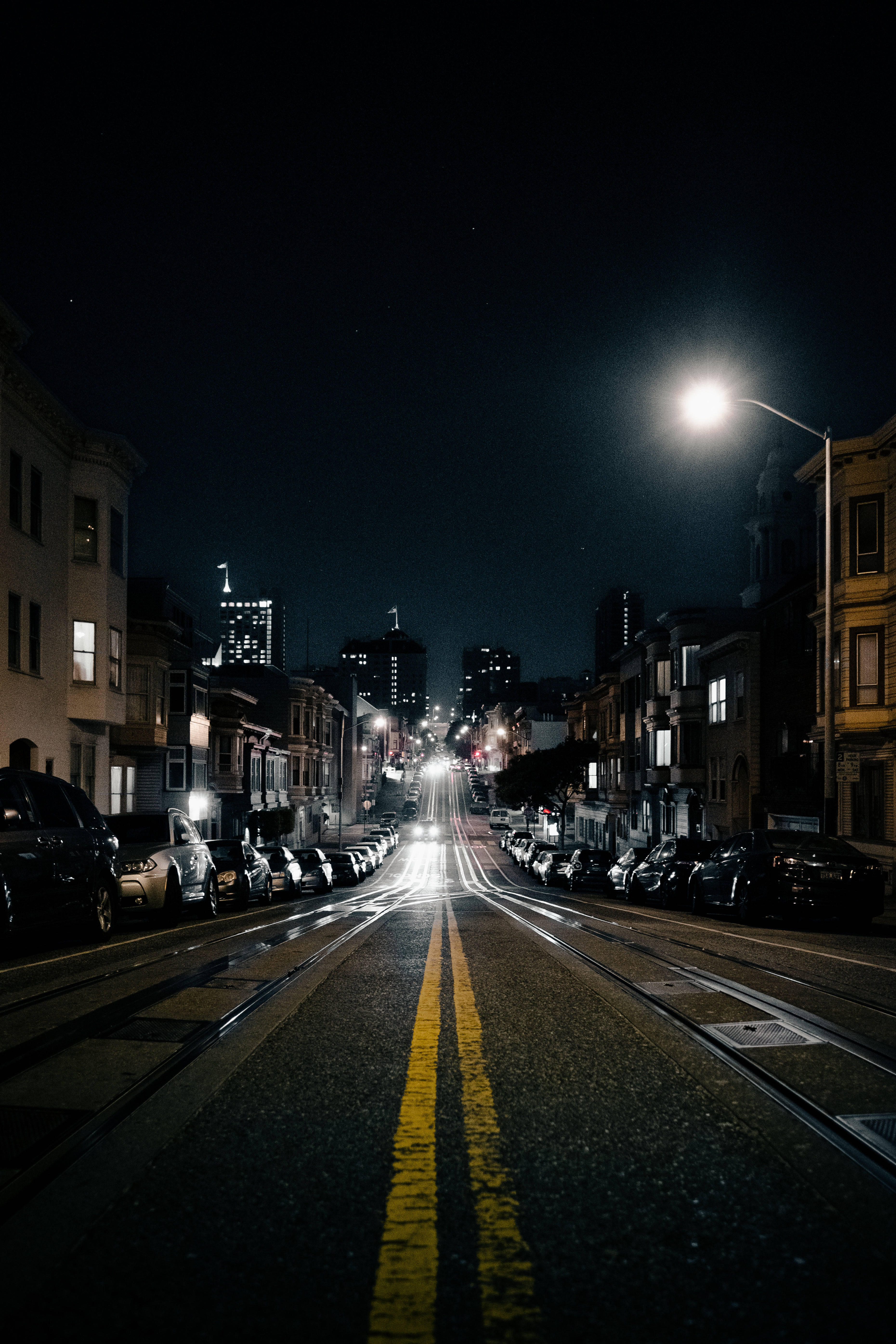 好看 桌布 壁紙 — 夜晚的城市街道，兩旁停滿汽車，發光的街燈延伸至遠處燈火通明的建築物。