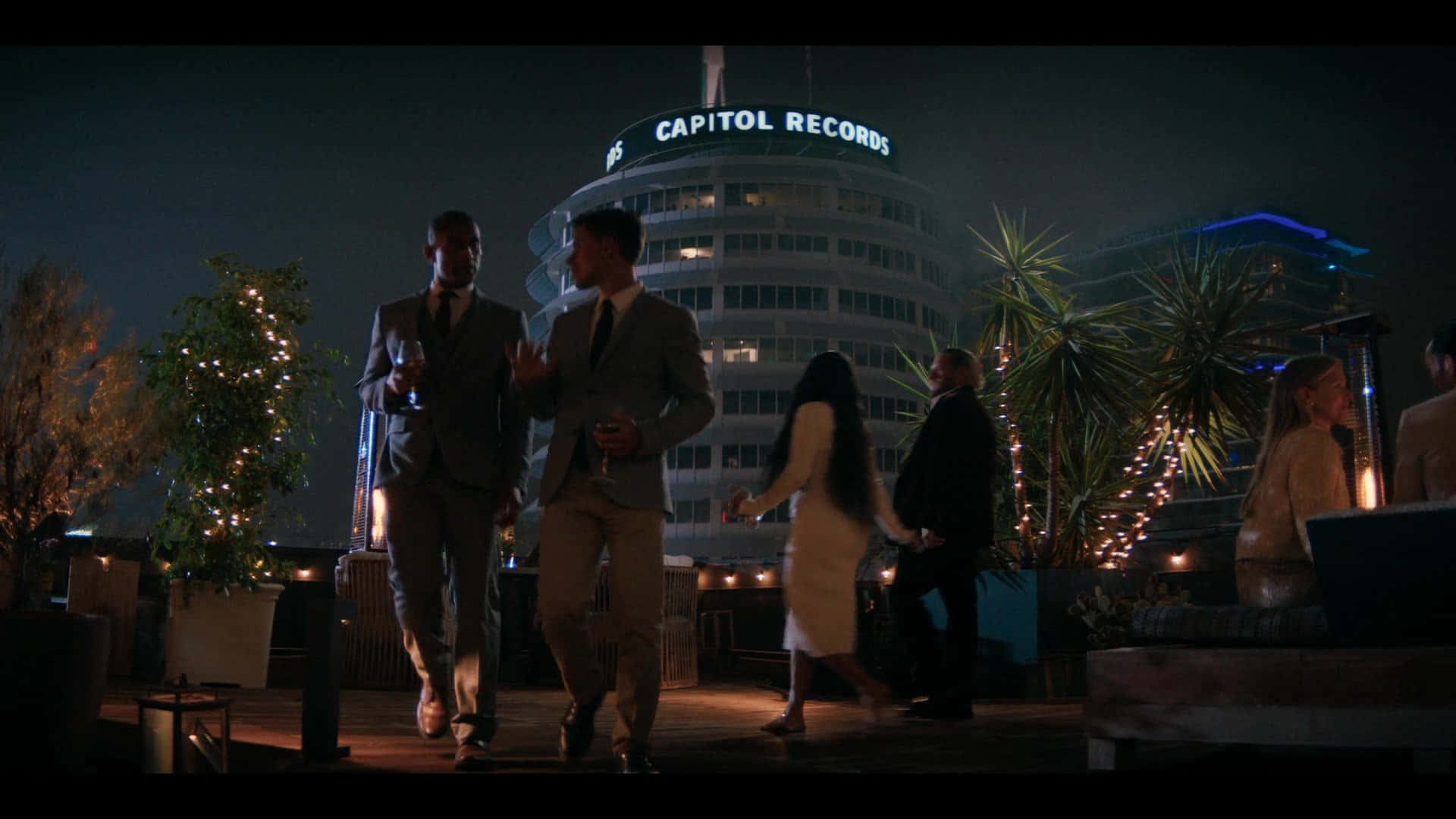 3D 桌布 壁紙 — 夜晚的屋頂派對，賓客在燈串下交流，背景是發光的Capitol Records大樓。