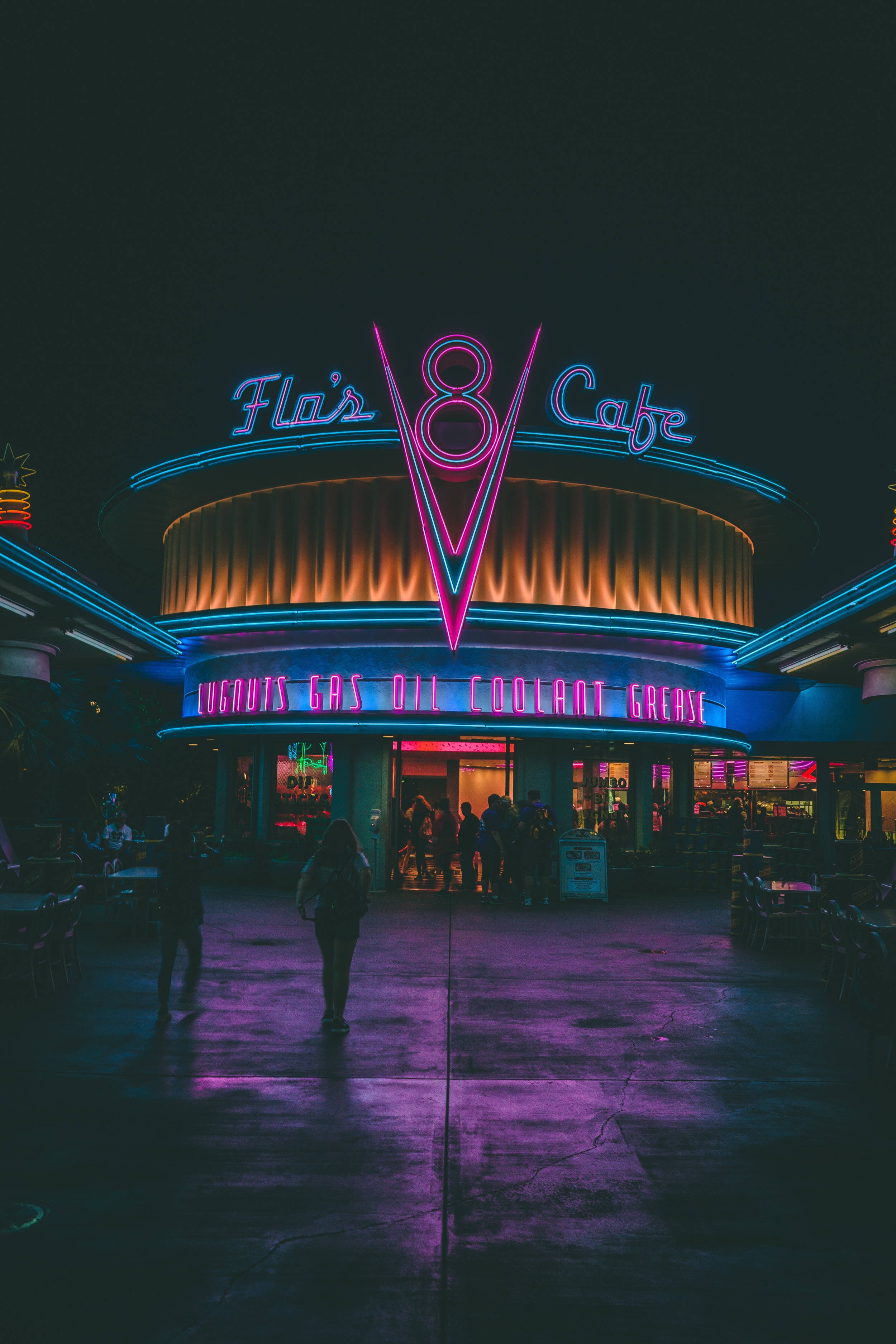 Disneyland 桌布 壁紙 — 夜晚的復古餐廳閃爍著鮮豔的霓虹燈，紫色和藍色的光線投射在濕漉漉的人行道上，剪影人物走過。