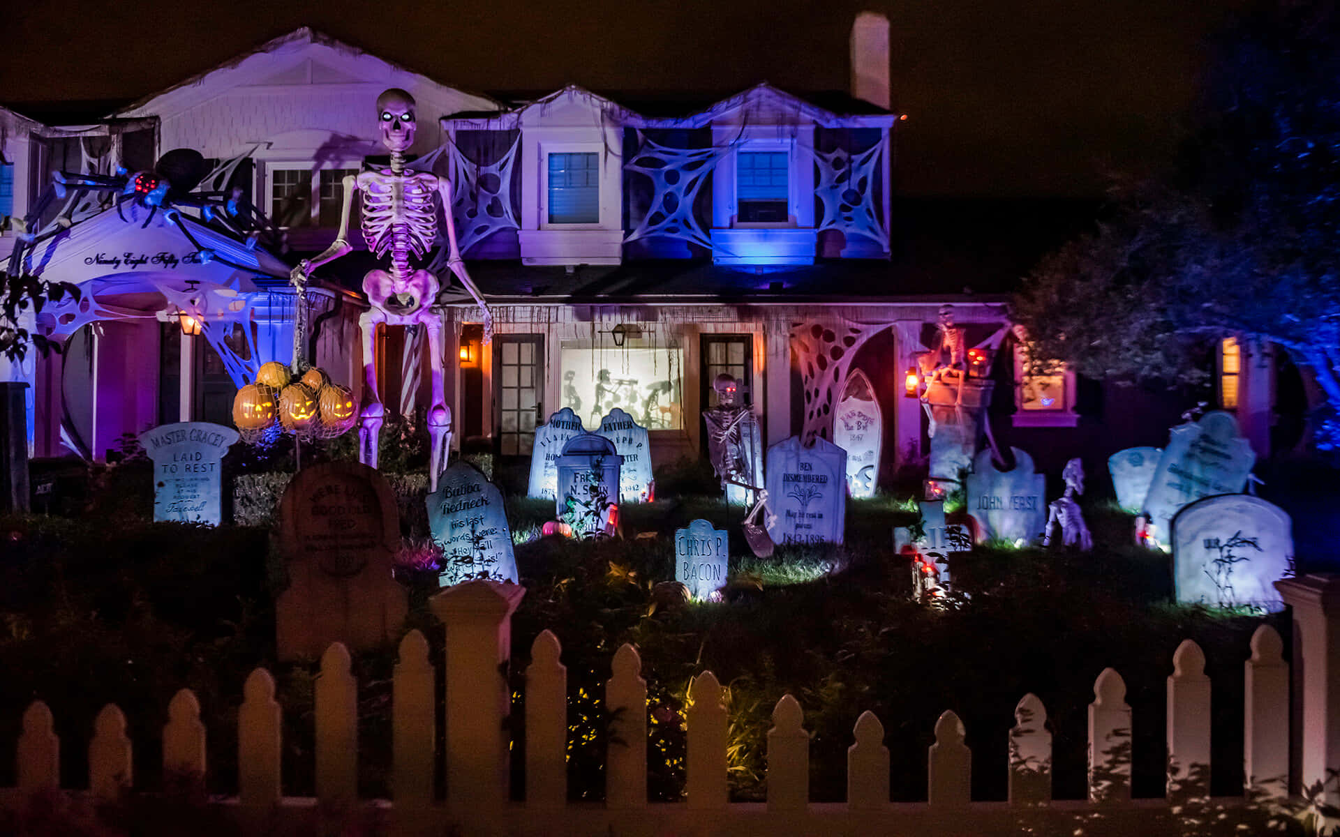 Halloween Yard Decorations 桌布 壁紙 — 夜晚的房子，裝飾著發光的傑克南瓜燈、骷髏和墓碑，在紫色和藍色燈光下。