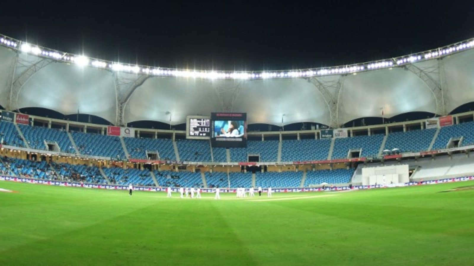 Cricket Ground 桌布 壁紙 — 夜晚的板球場，明亮的泛光燈照亮綠色球場和藍色座位，天空漆黑。