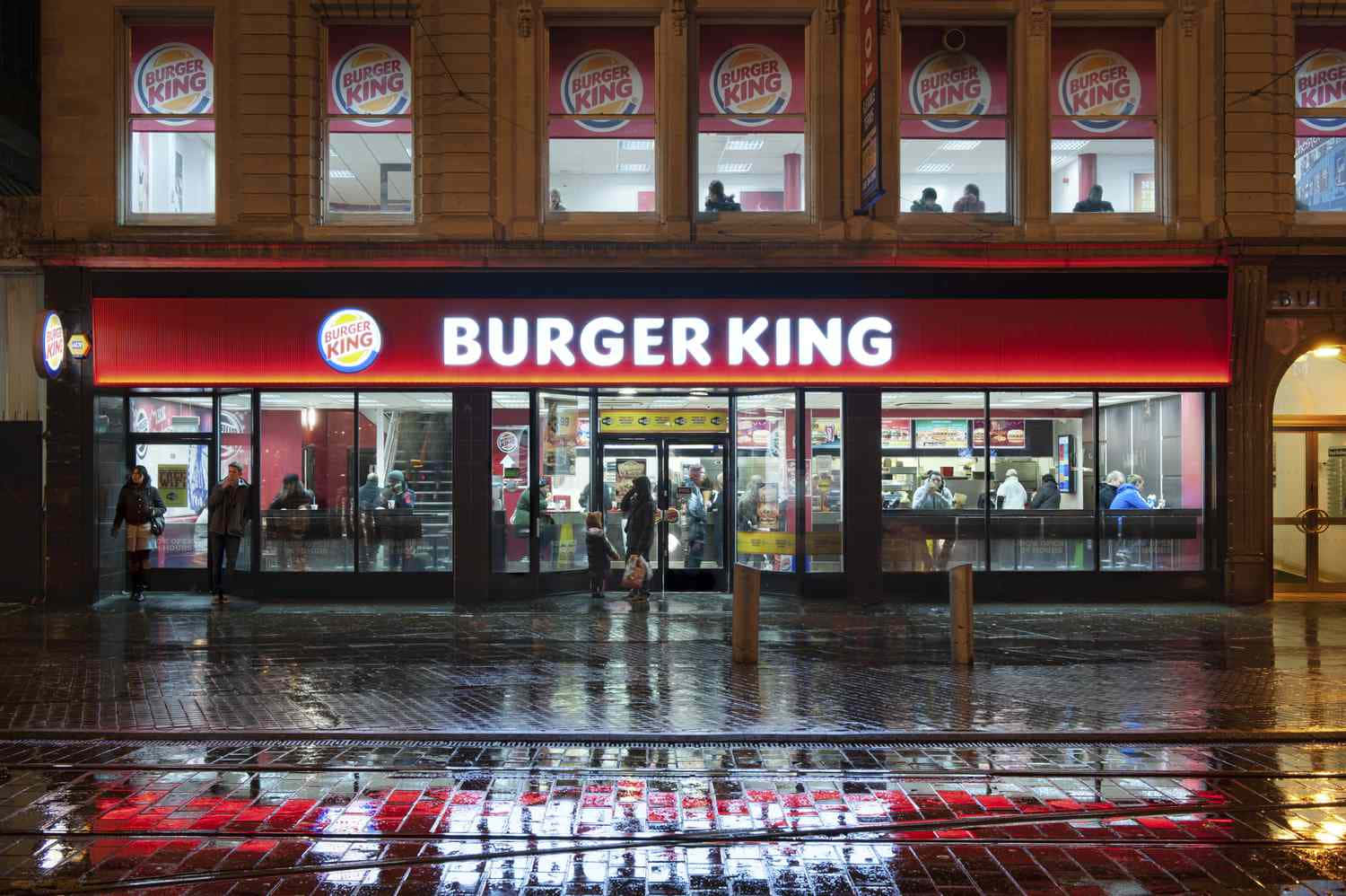 Burger King 桌布 壁紙 — 夜晚的漢堡王餐廳，濕漉漉的路面反射出其明亮的紅色霓虹燈招牌和顯示店內顧客的窗戶。