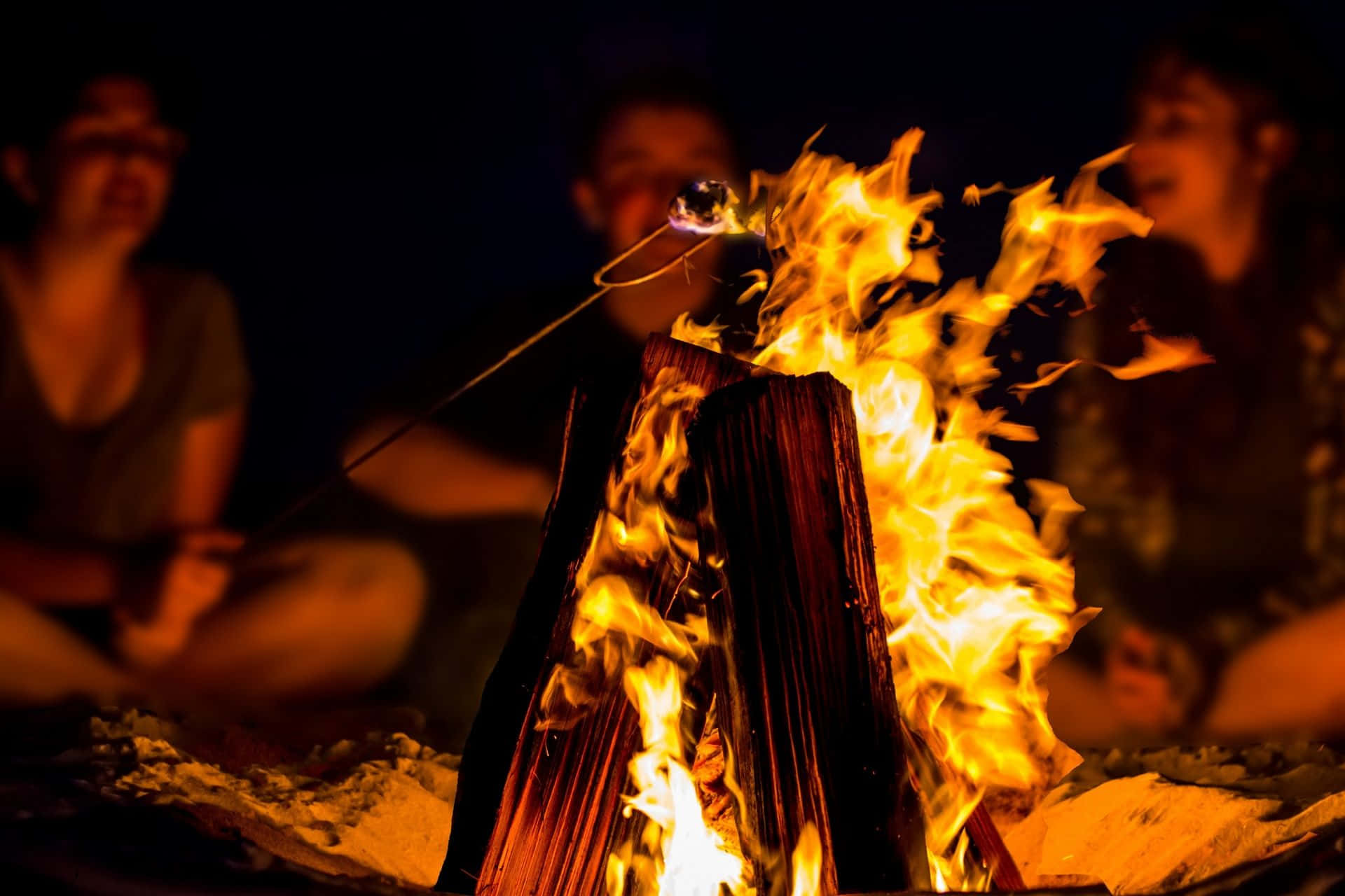 Beach Bonfire 桌布 壁紙 — 夜晚的營火咆哮，人們正在烤棉花糖，發光的餘燼將溫暖的光投射在他們的臉上。