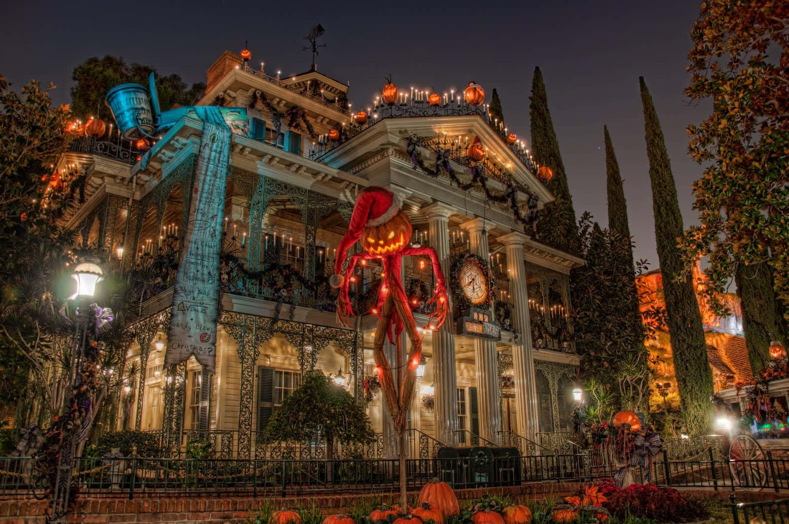 Disney Halloween 桌布 壁紙 — 夜晚的維多利亞式大宅，裝飾著發光的南瓜燈和前面一個巨型南瓜頭稻草人。