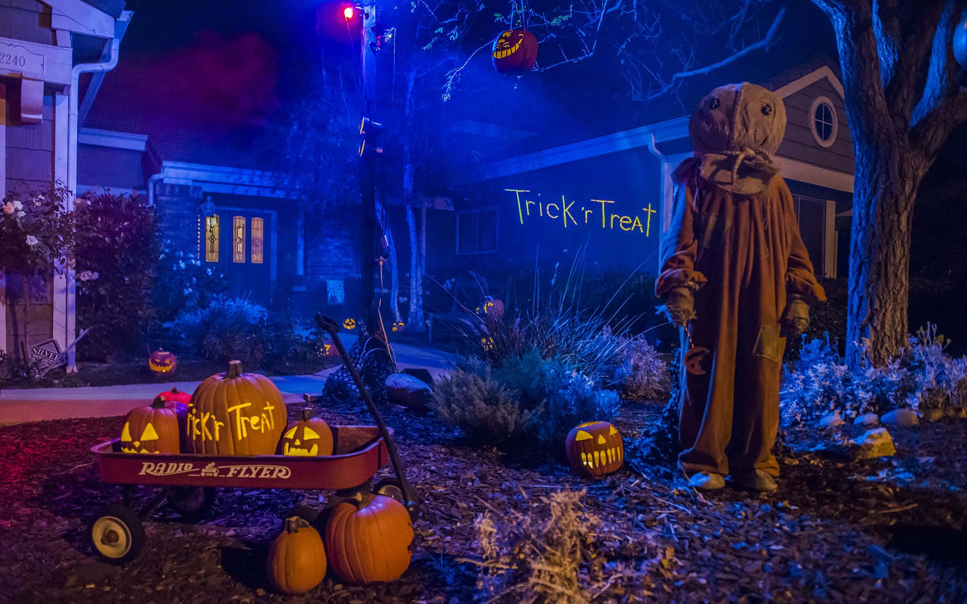 Trick Or Treat 桌布 壁紙 — 夜晚的詭異萬聖節場景，有稻草人、發光的傑克南瓜燈和寫著「不給糖就搗蛋」的馬車，籠罩在怪異的藍光下。