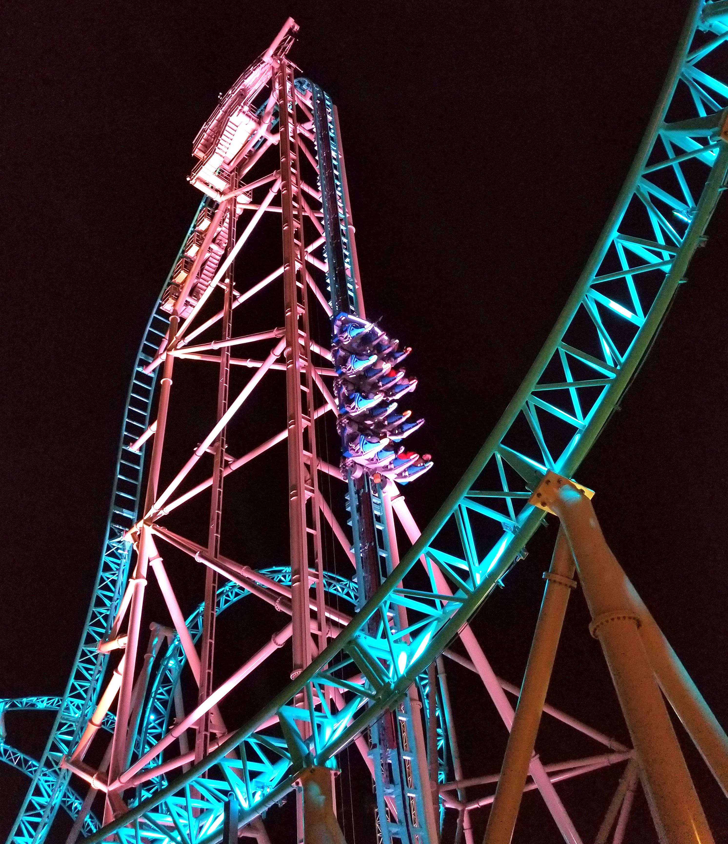 Roller Coaster 桌布 壁紙 — 夜晚的雲霄飛車，乘客正攀爬一座陡峭的粉紅色照明塔，背景是黑暗的天空。
