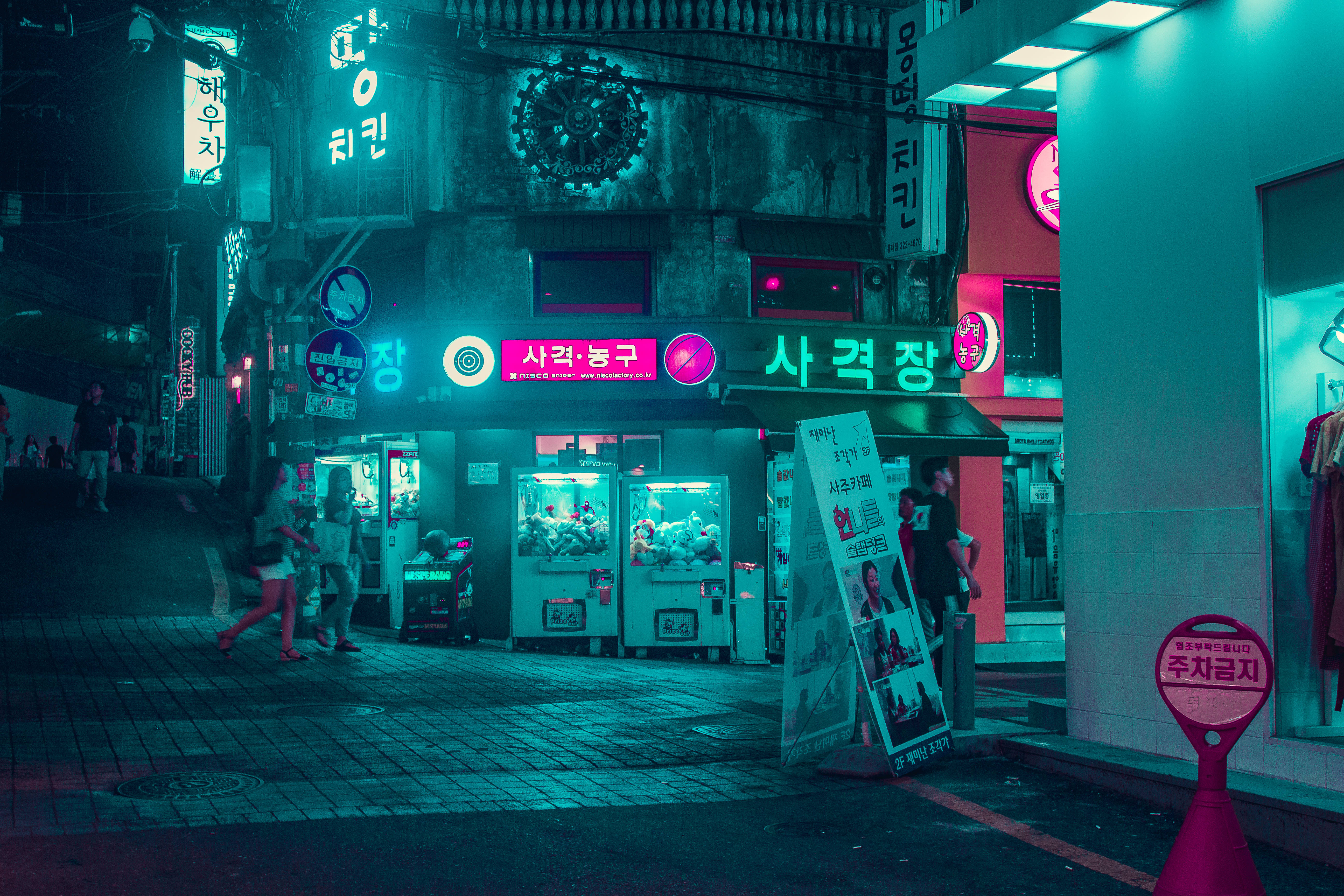 cyberpunk ultrawide 桌布 壁紙 — 夜晚的韓國小巷，霓虹燈閃爍，有發光招牌、夾娃娃機和行人走在濕漉漉的鵝卵石街道上。