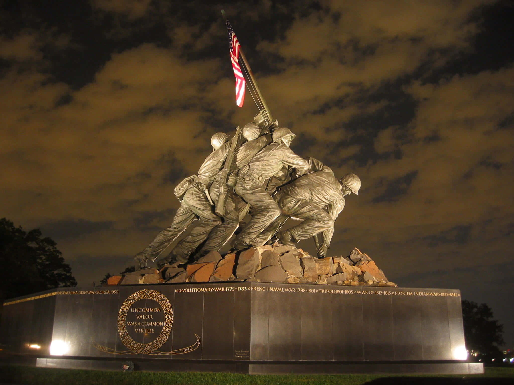 US Marines 桌布 壁紙 — 夜晚紀念碑，士兵在岩石底座上舉旗，天空多雲，配以溫暖的人造燈光。
