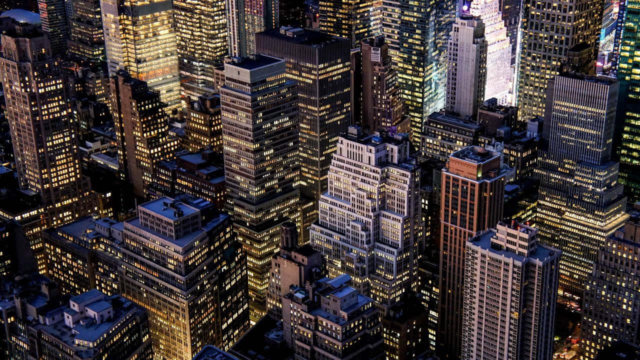 720p Empire State Building 桌布 壁紙 — 夜晚繁忙城市中高聳摩天大樓的鳥瞰圖，帶有發光的窗戶和密集的城市建築。