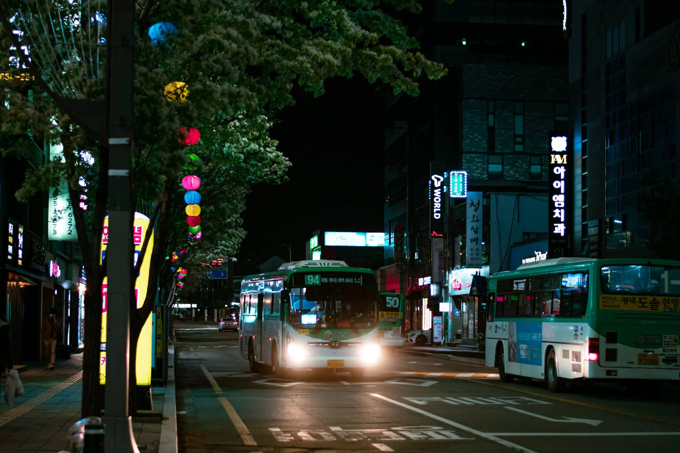 城市 桌布 壁紙 — 夜晚街景，有發光的霓虹燈招牌和兩輛綠色公車，樹枝下掛著彩色燈籠。