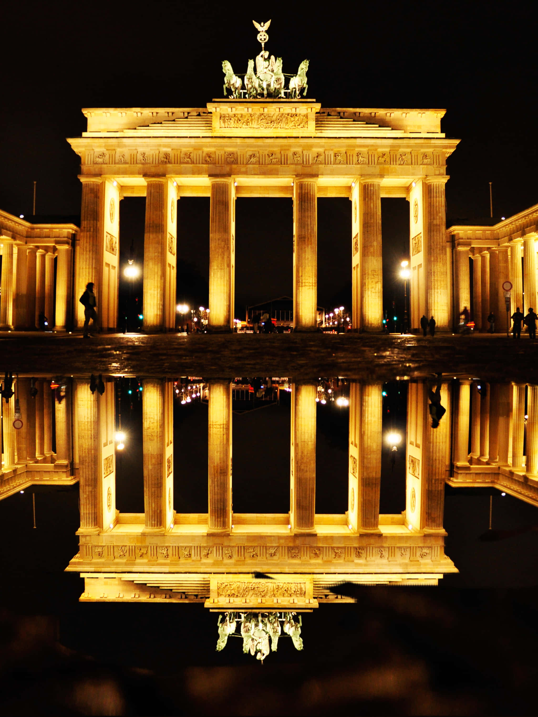 Berlin 桌布 壁紙 — 夜晚被照亮的勃蘭登堡門在濕漉漉的路面上完美倒映，形成對稱的金色鏡面效果。