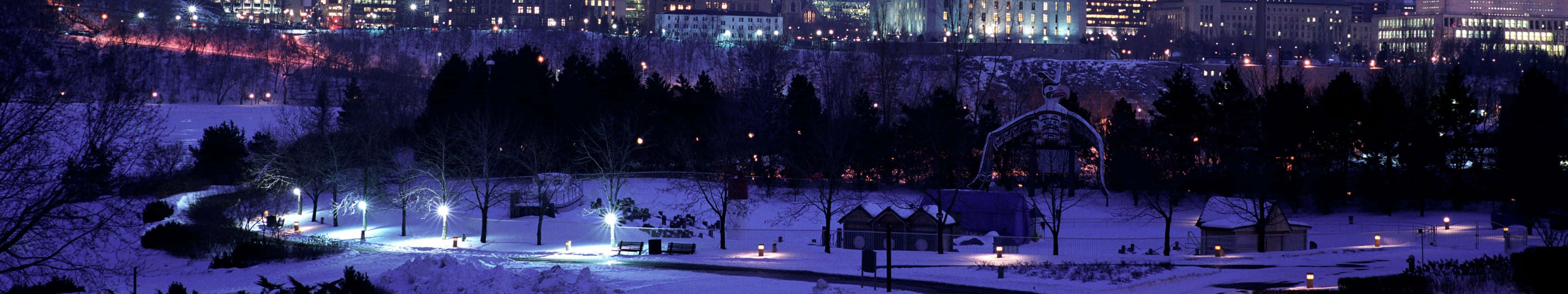 4098x768 桌布 壁紙 — 夜晚被發光燈光照亮的雪地公園，背景中城市天際線閃爍。