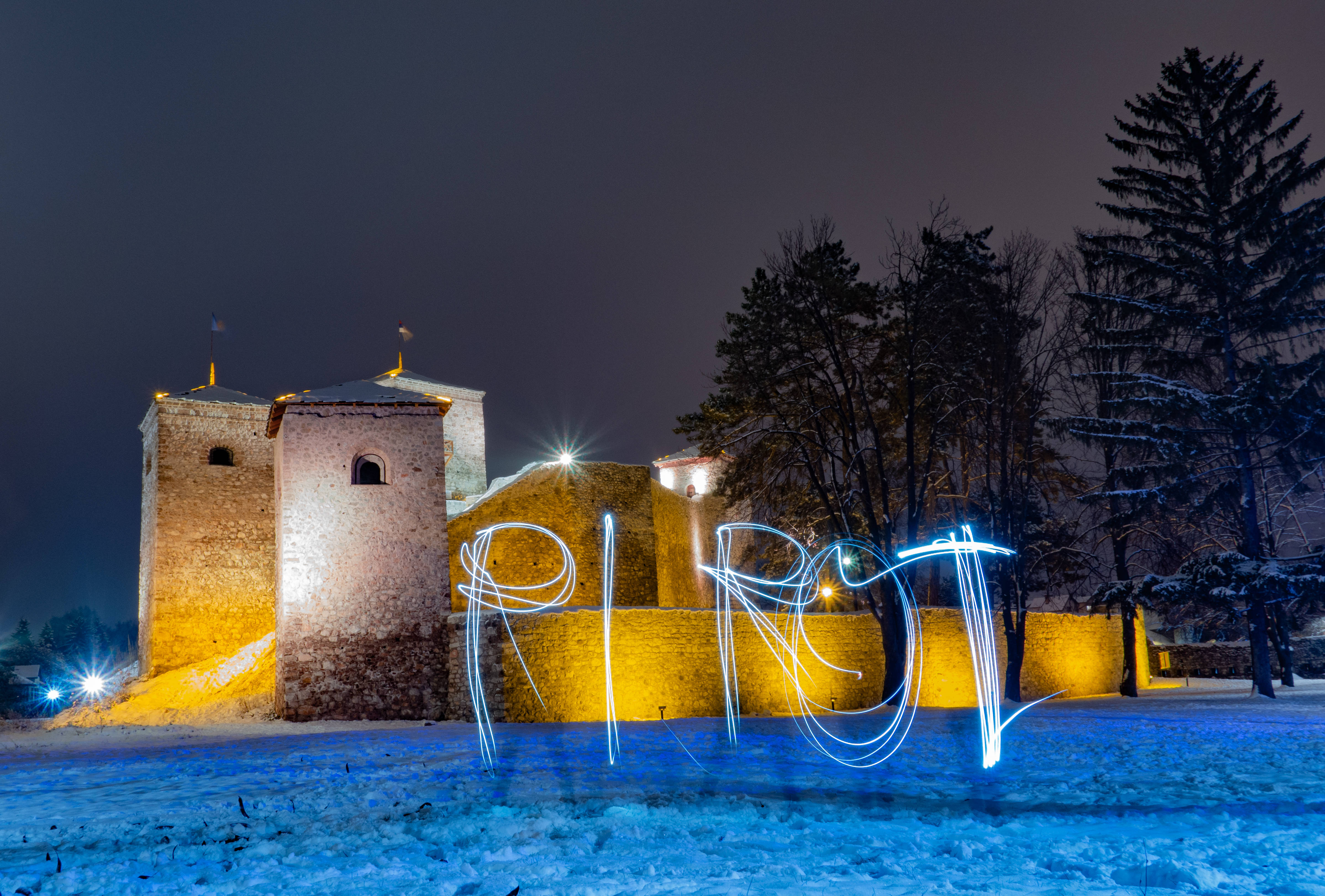 Serbia 桌布 壁紙 — 夜晚被雪覆蓋的中世紀堡壘，被溫暖的燈光照亮，前景中有發光的藍色光繪字母拼出「PIROT」。