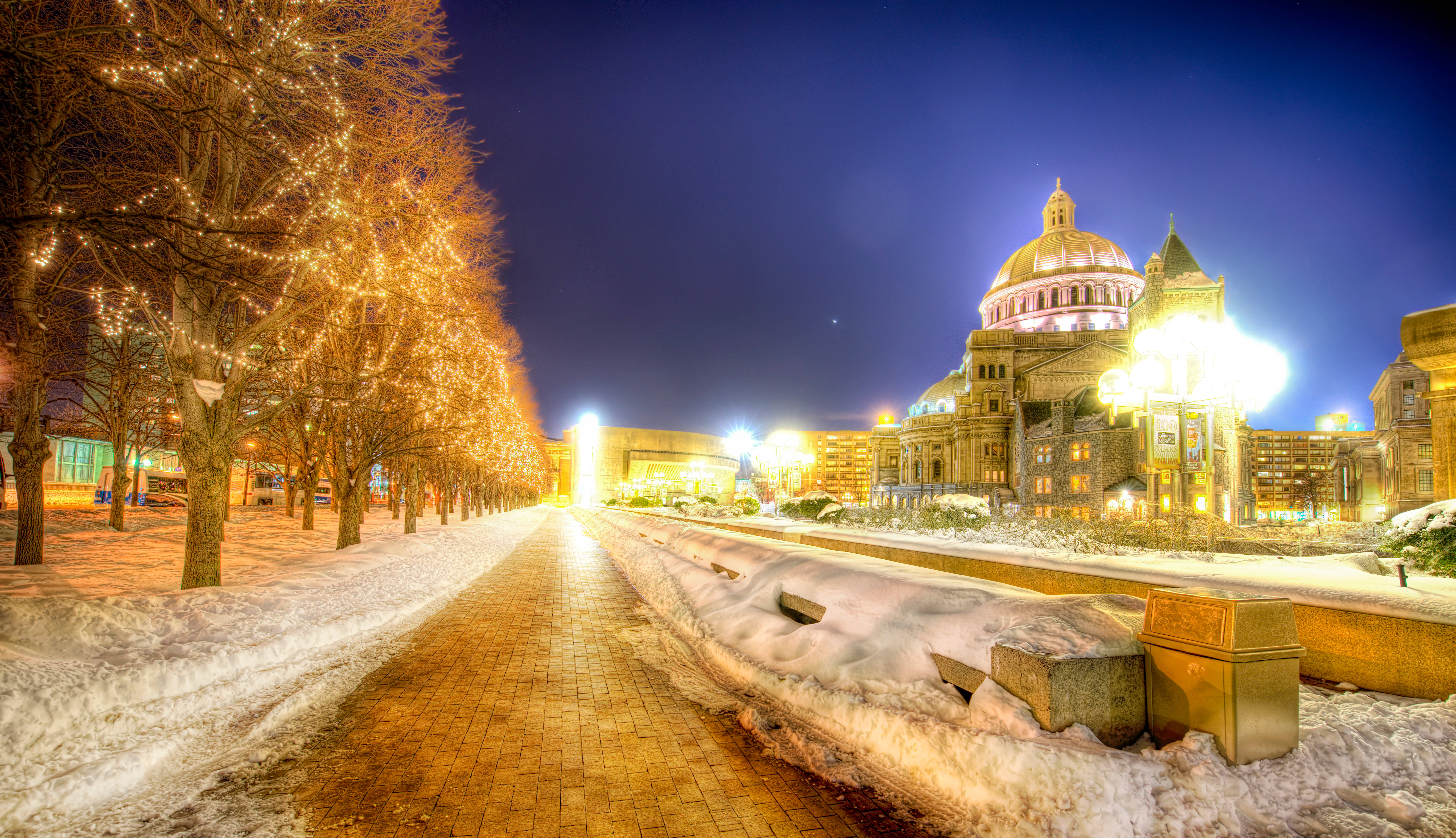 Boston 桌布 壁紙 — 夜晚被雪覆蓋的城市街道，兩旁光禿禿的樹木上掛滿了發光的燈串，通向一座宏偉的燈光大教堂。