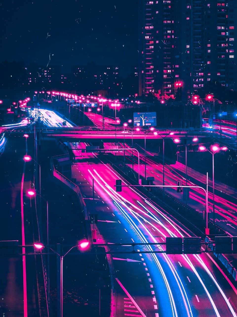 桌布 壁紙 — 夜晚被霓虹燈淹沒的城市高速公路，高聳摩天大樓下行駛的汽車留下粉紅色和藍色的光跡。