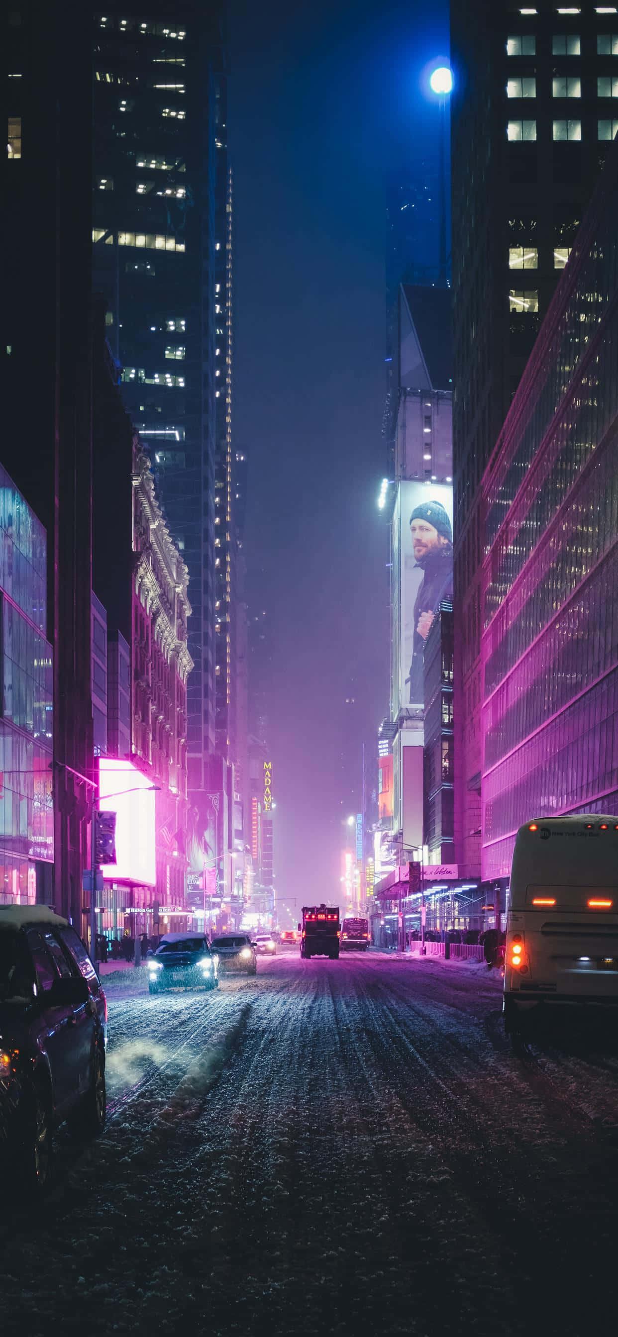 New York City Night iPhone 桌布 壁紙 — 夜晚被高聳摩天大樓和發光廣告牌散發的鮮豔粉紅和藍色霓虹燈光沐浴的積雪城市街道。