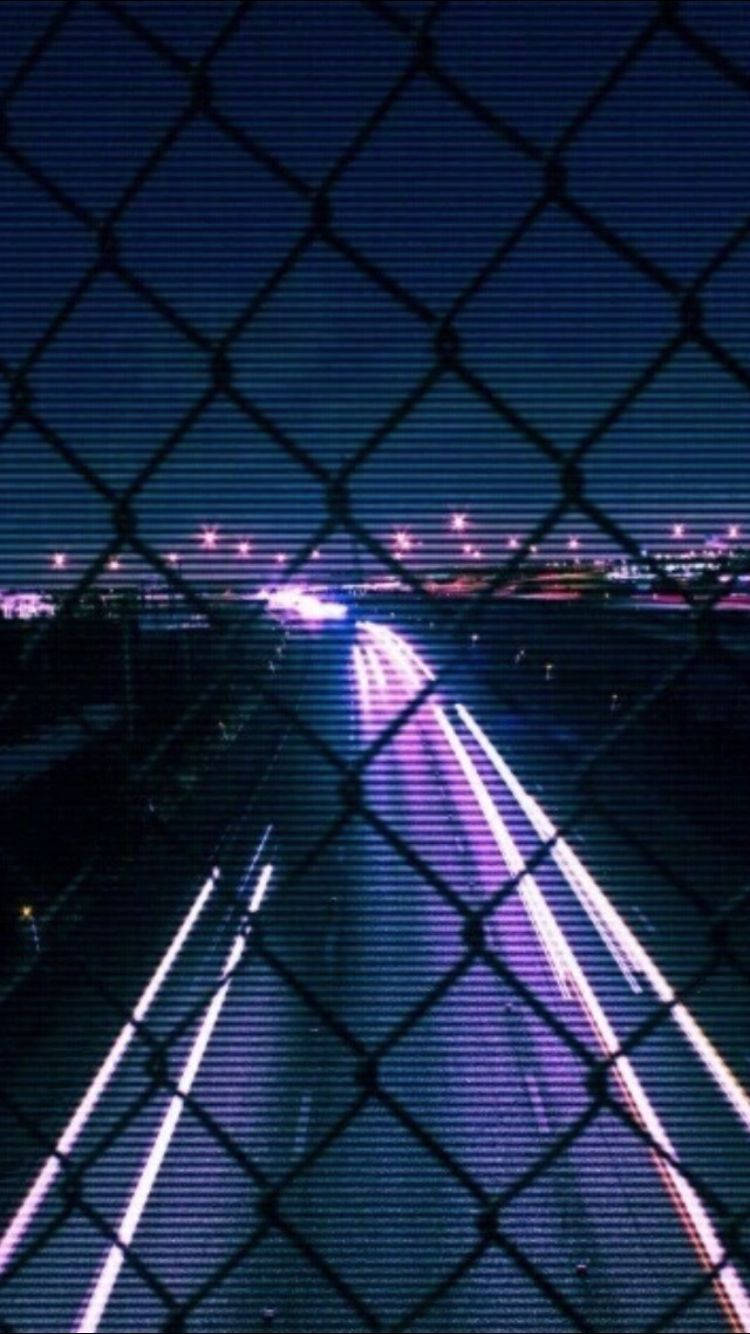 黑紫美學 桌布 壁紙 — 夜晚透過鐵絲網看到的霓虹高速公路，帶有紫色和白色光的發光條紋。