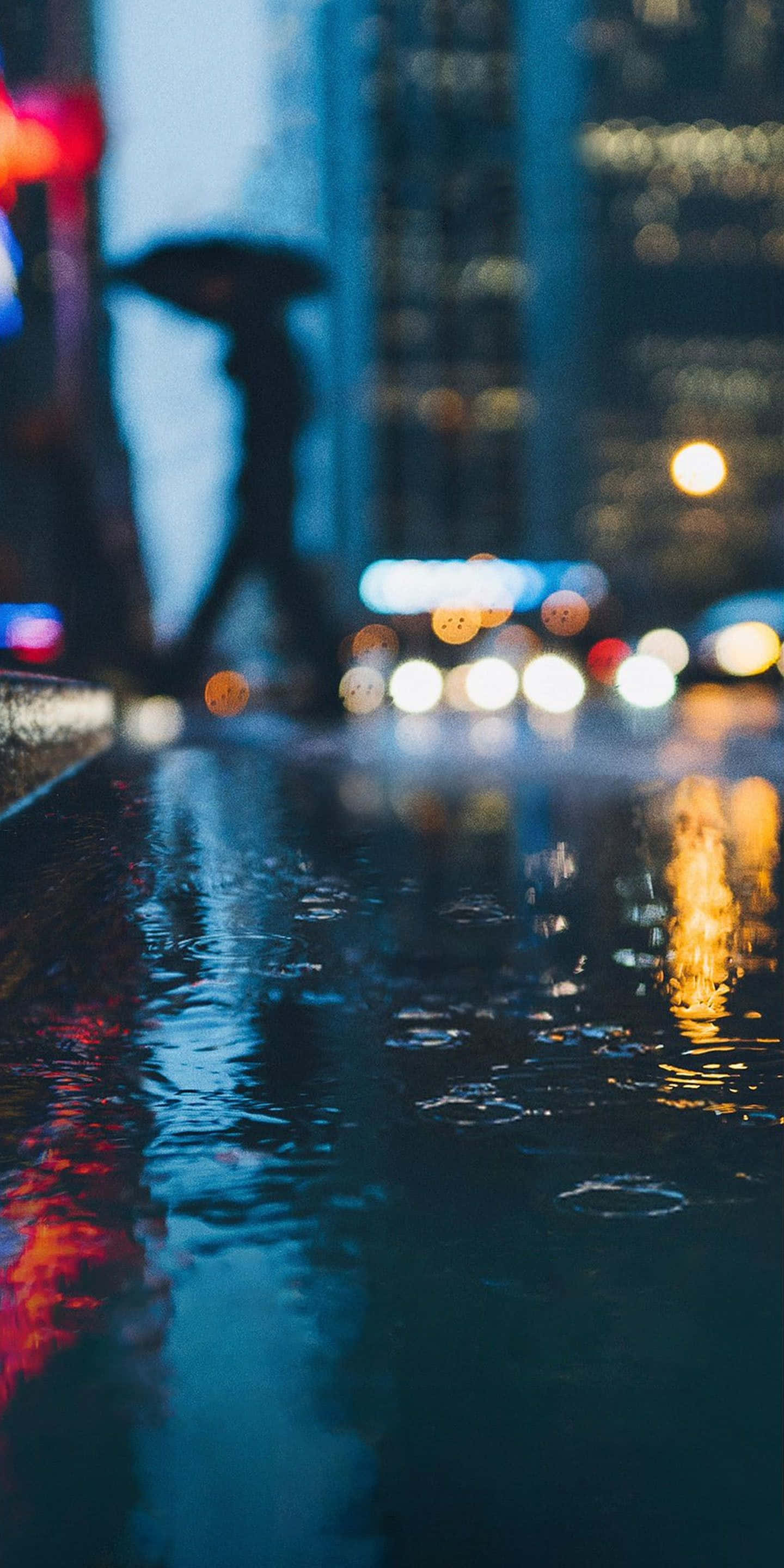 桌布 壁紙 — 夜晚雨中的城市街道，模糊的城市燈光在濕漉漉的路面上反射，一個撐著雨傘的剪影人物。
