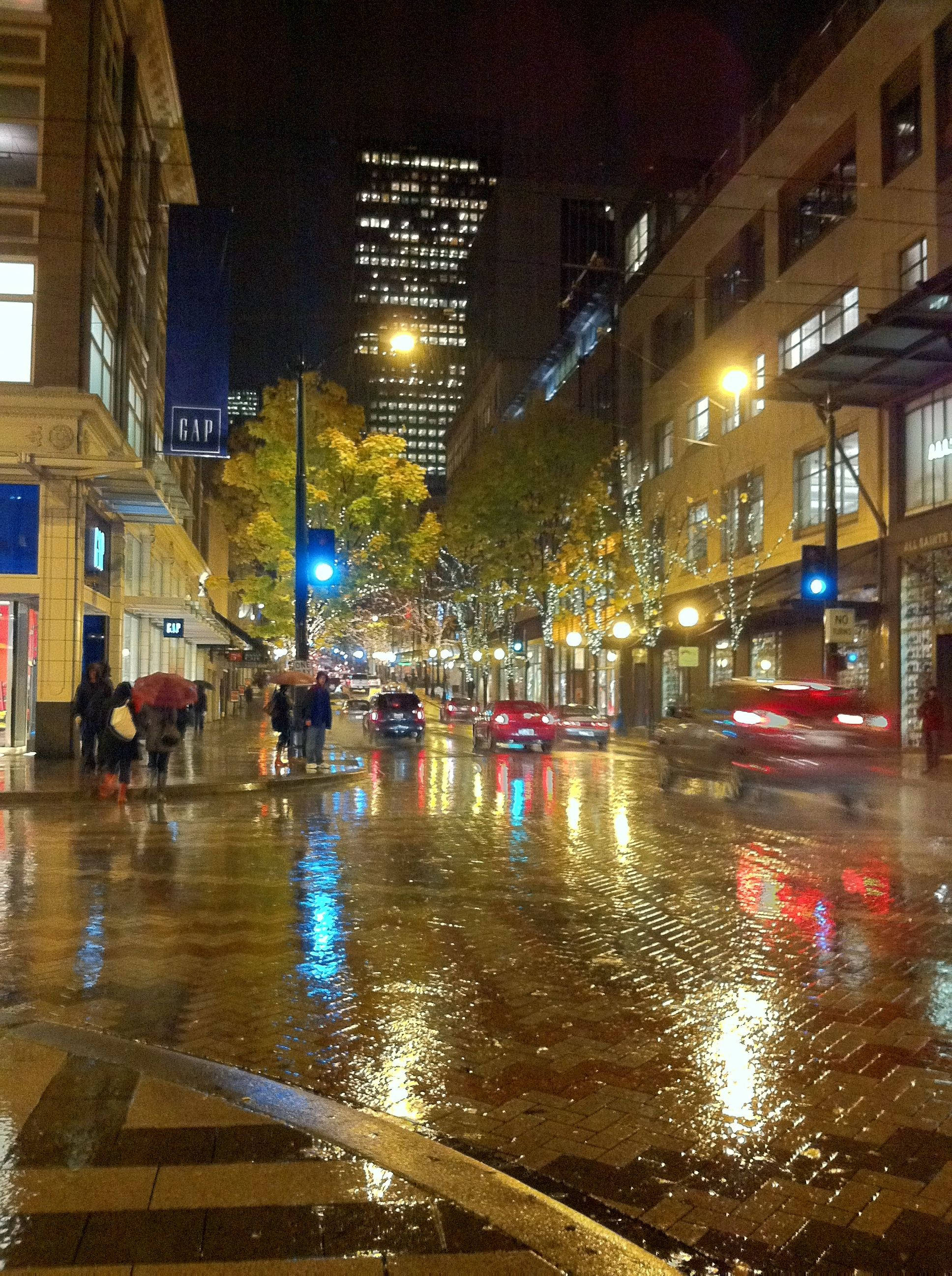 Rainy Day 桌布 壁紙 — 夜晚雨濕的城市街道，濕漉漉的路面反射著閃爍的路燈，行人撐著雨傘。