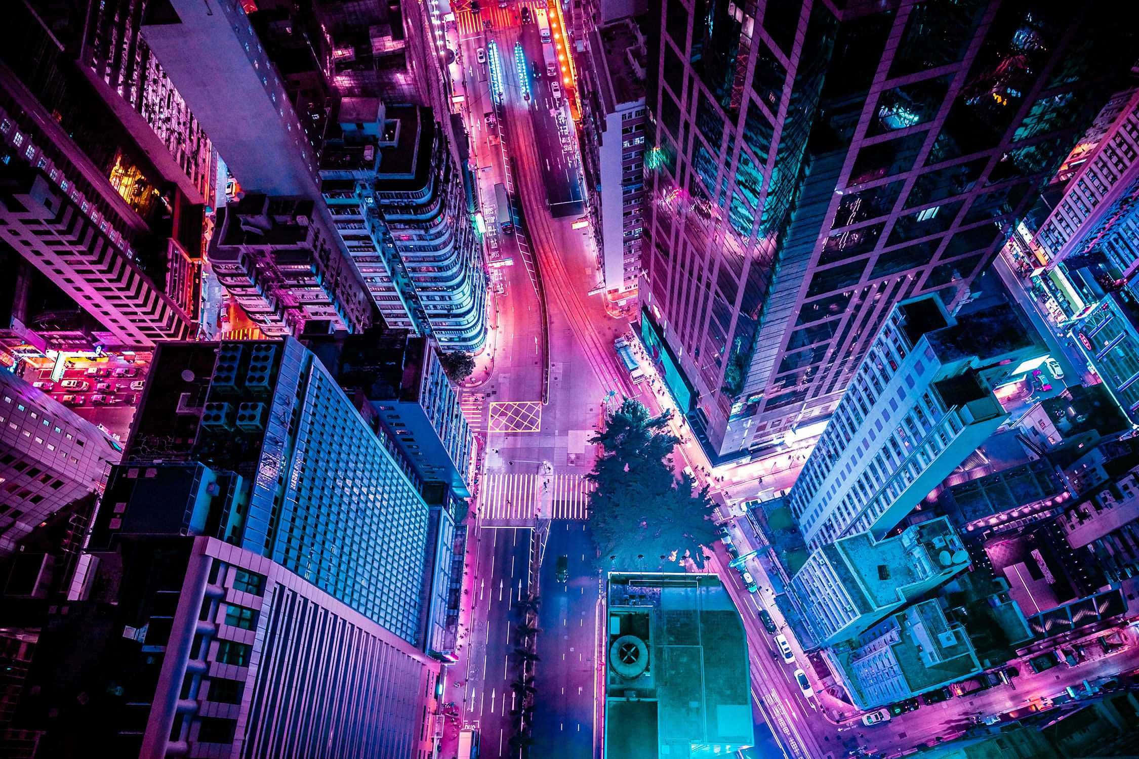 美學 桌布 壁紙 — 夜晚霓虹燈光璀璨的城市街道鳥瞰圖，高聳的摩天大樓閃爍著電光紫和藍色調。