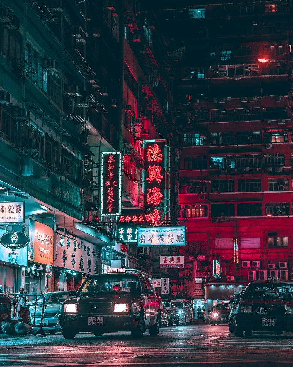 桌布 壁紙 — 夜晚霓虹燈照亮嘅城市小巷，紅色同藍色嘅招牌喺濕漉漉嘅行人路上反射，的士駛過。