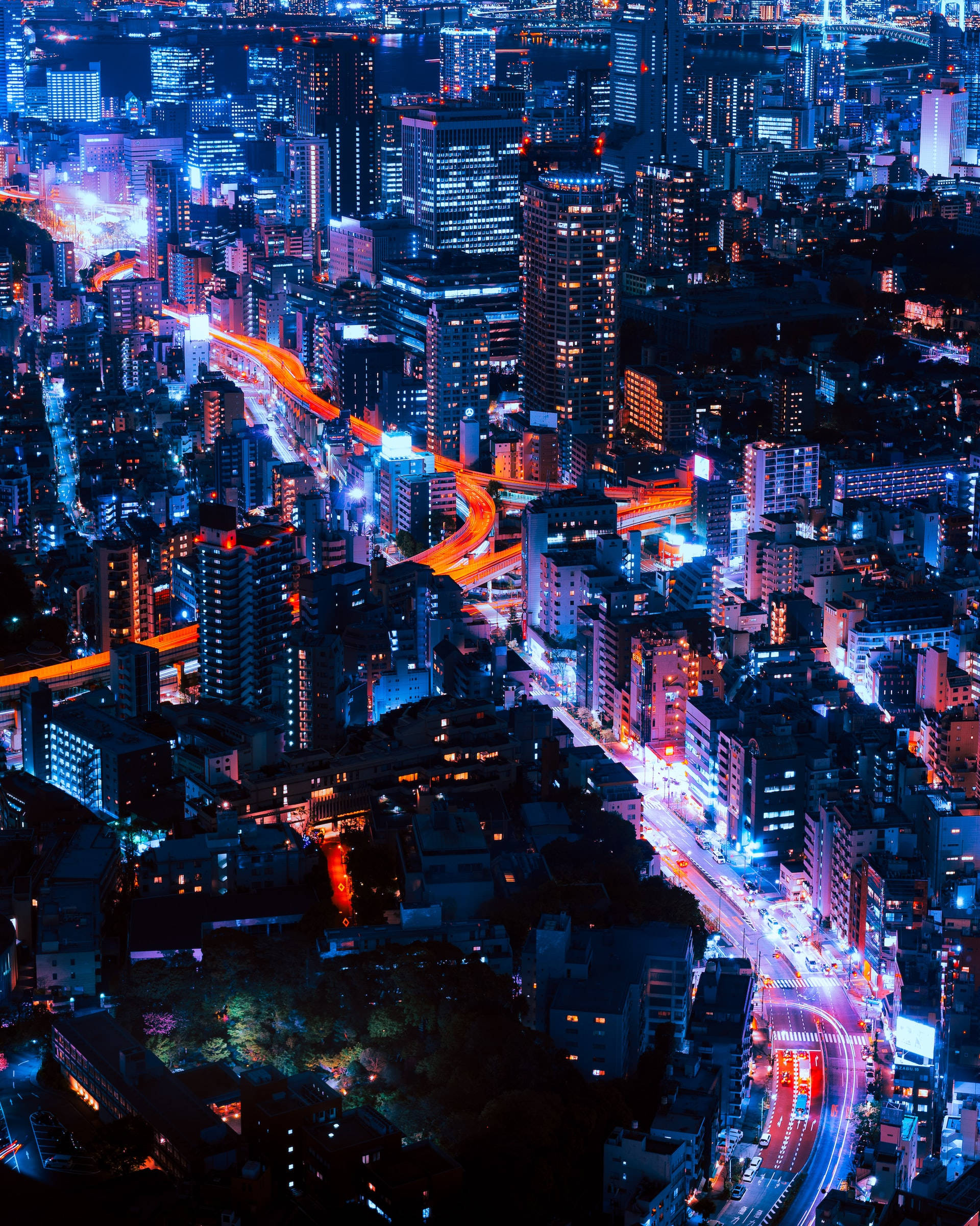 Skyscraper 桌布 壁紙 — 夜晚霓虹燈照亮城市景觀的航拍圖，建築物和交通痕跡散發出藍色和橙色的光芒。