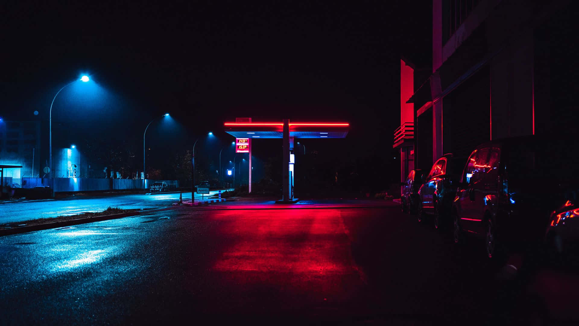 夜景城市 桌布 壁紙 — 夜晚霓虹燈照亮的加油站，紅色和藍色的燈光反射在潮濕的路面和停放的汽車上。