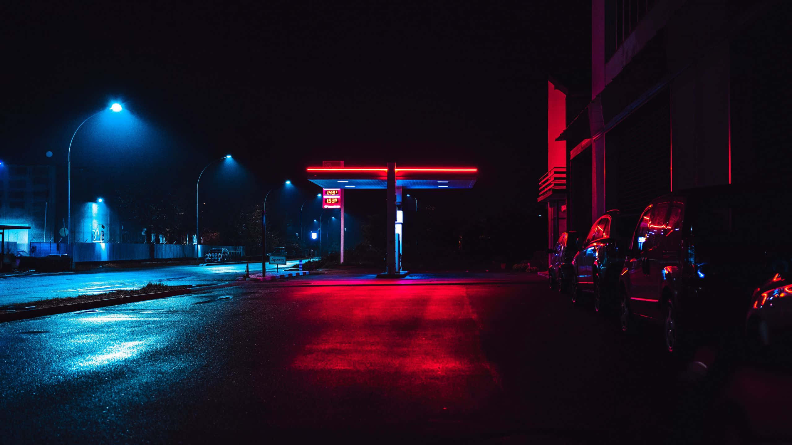 霓虹城市動態圖 桌布 壁紙 — 夜晚霓虹燈照亮的加油站，紅色和藍色的燈光反射在濕漉漉的路面和停泊的汽車上。