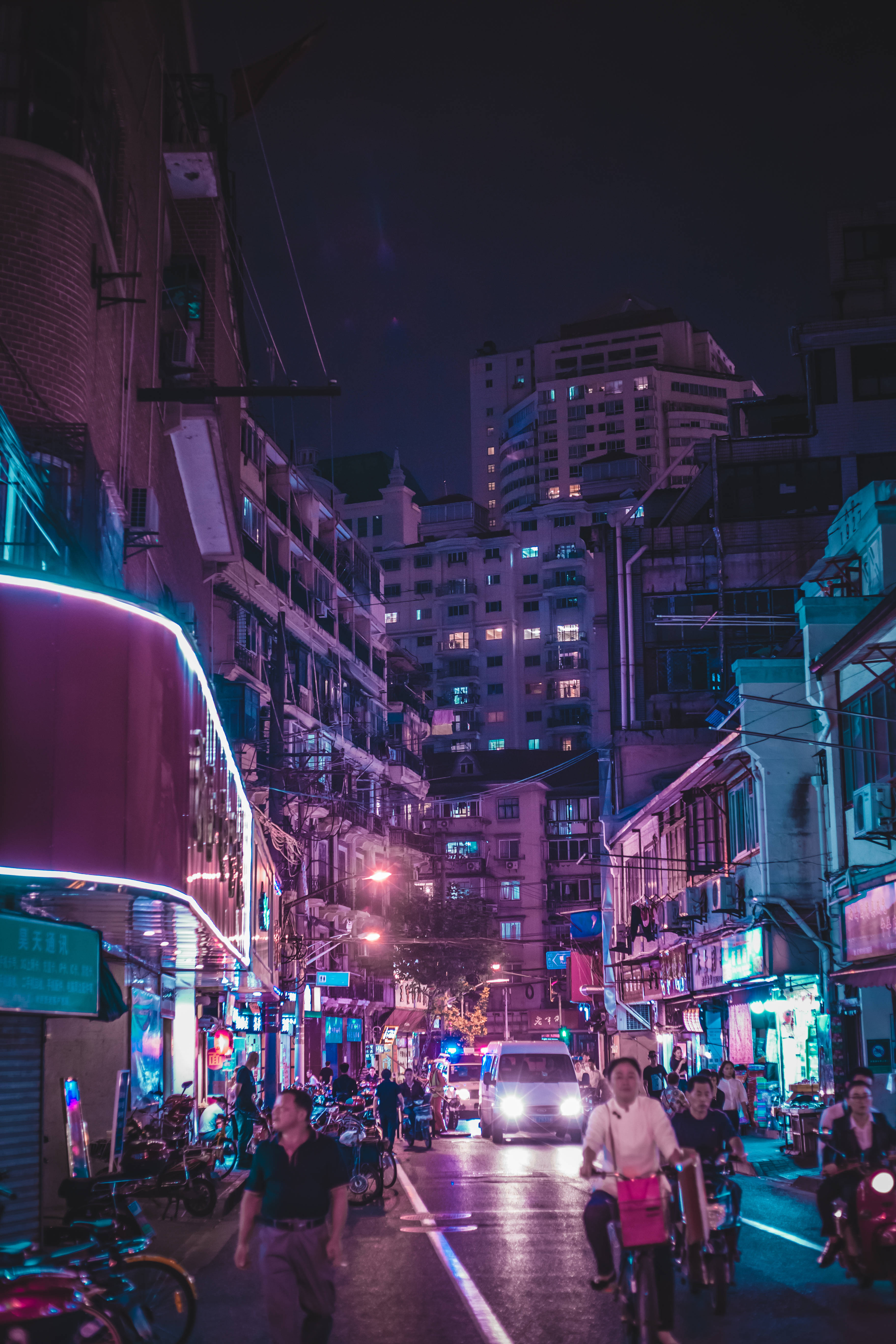 淡紫色 桌布 壁紙 — 夜晚霓虹燈照亮的城市小巷，人們騎著腳踏車和步行經過發光的商店櫥窗和高聳的公寓大樓。
