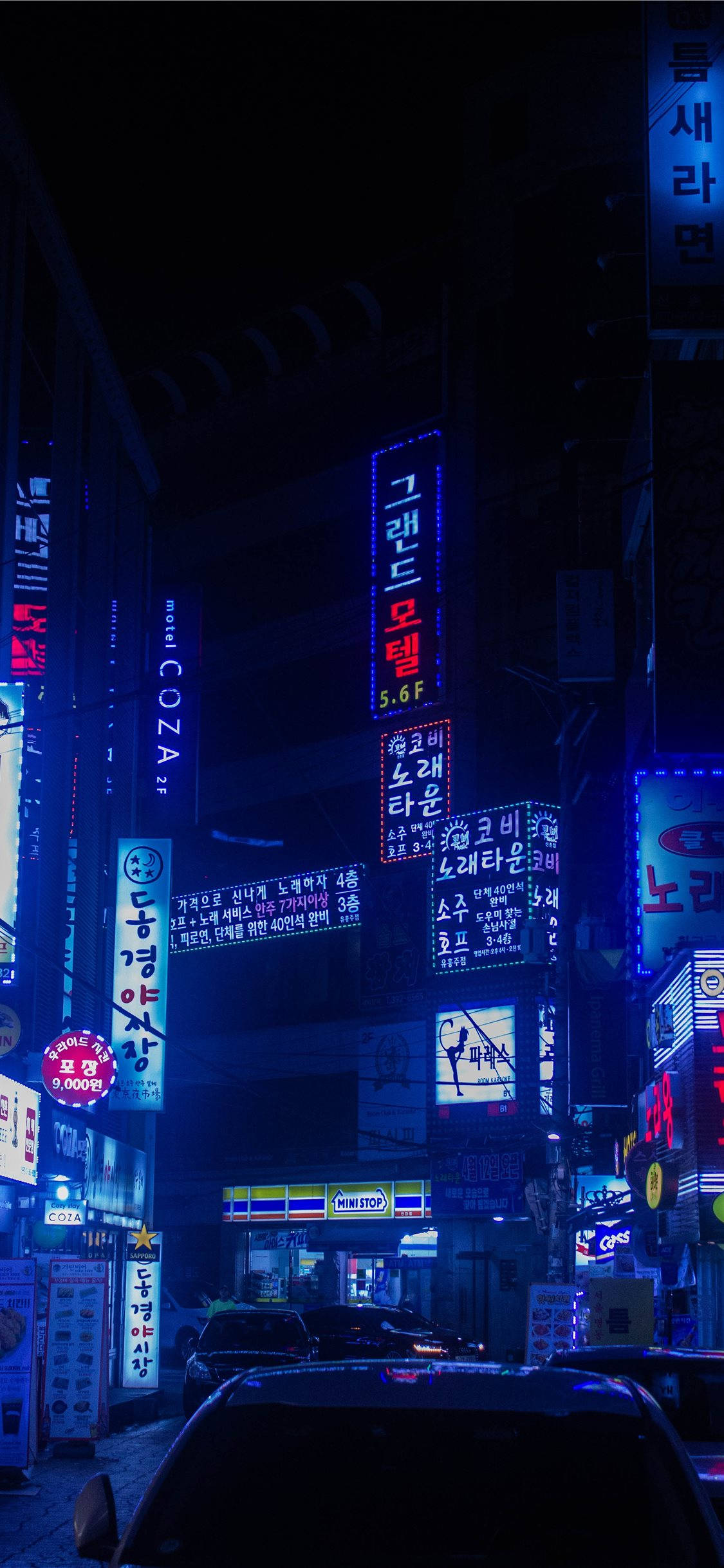 手機 桌布 壁紙 — 夜晚霓虹燈照亮的城市小巷，發光的韓文招牌倒映在濕漉漉的人行道和停放的汽車上。