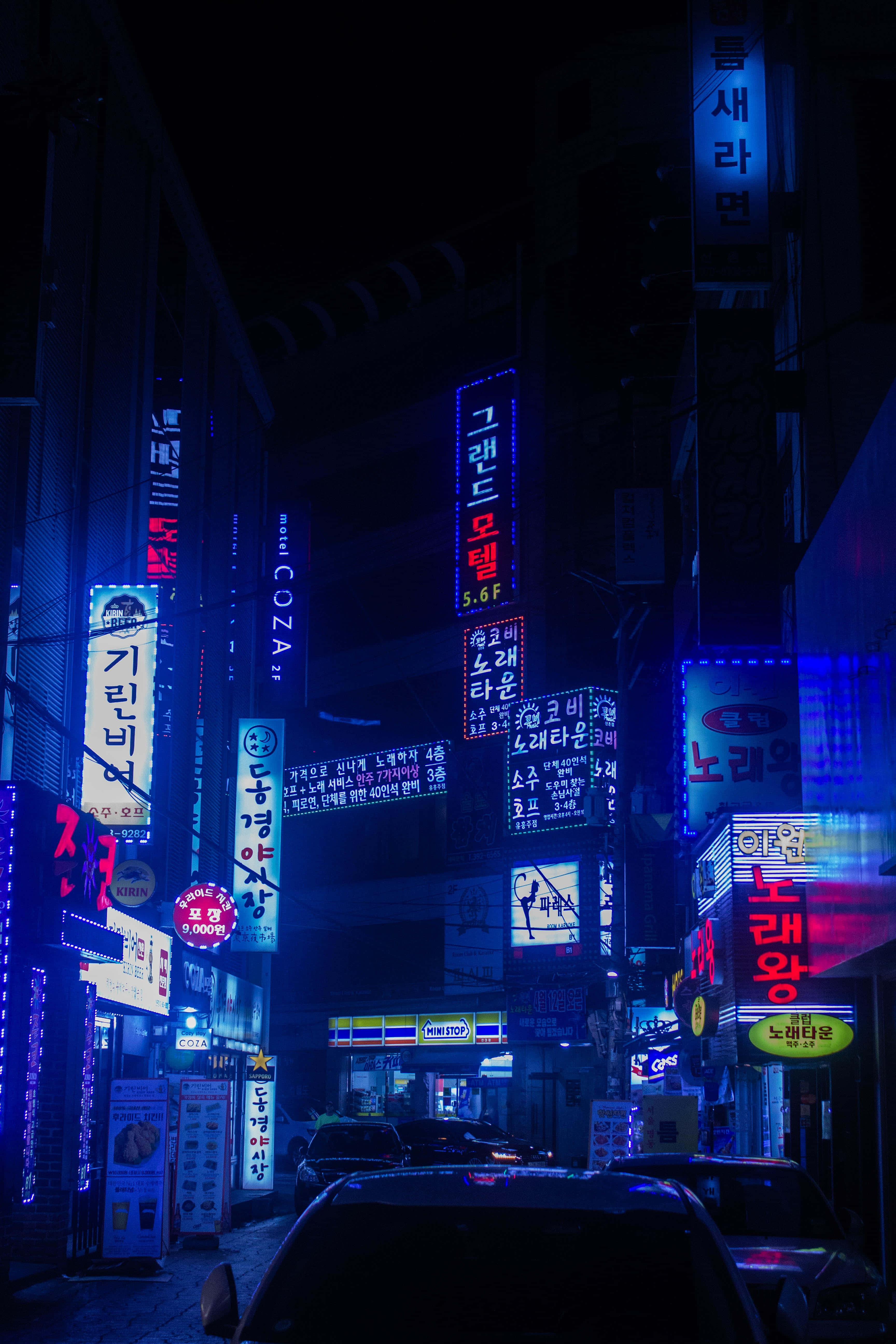 韓國 桌布 壁紙 — 夜晚霓虹燈照亮的城市小巷，韓文發光招牌在停泊的汽車和建築物上投射出電藍色和紅色色調。
