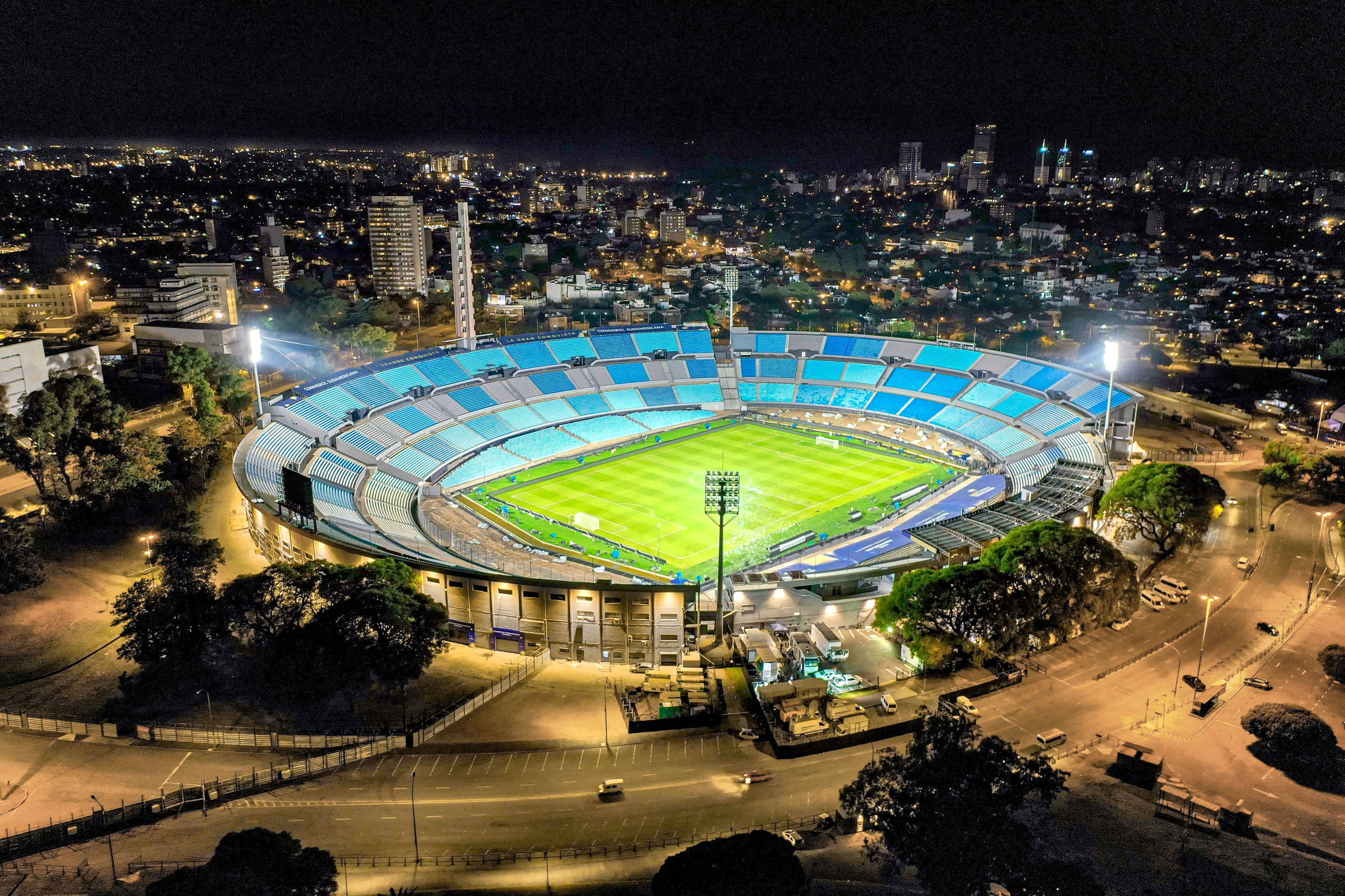 Stadium 桌布 壁紙 — 夜晚鳥瞰一個燈火通明的體育場，擁有發光的藍色座位和充滿活力的綠色場地，周圍環繞著城市燈光。