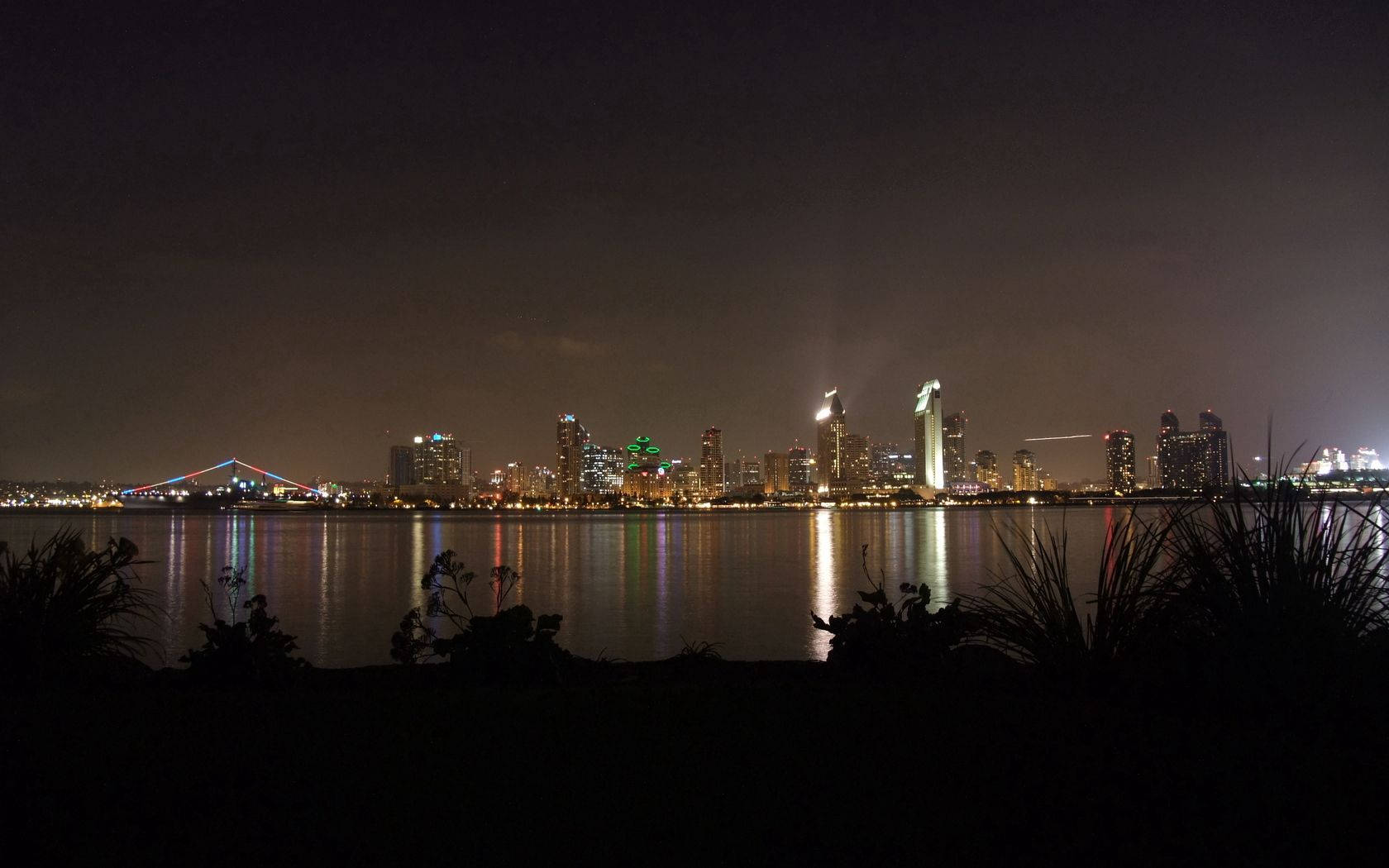 Beautiful Night 桌布 壁紙 — 夜景城市景觀，有燈火通明的摩天大樓和色彩繽紛的橋樑，倒映在平靜的水面上，周圍環繞著深色樹葉。