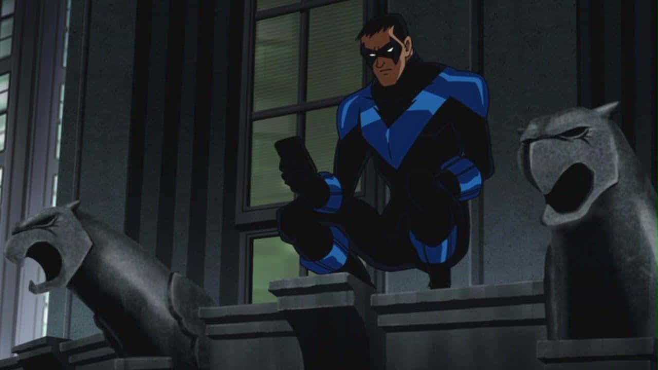 Batman Under The Red Hood 桌布 壁紙 — 夜翼在昏暗燈光下警覺地棲息在石像鬼上，背景是黑暗的城市建築立面。
