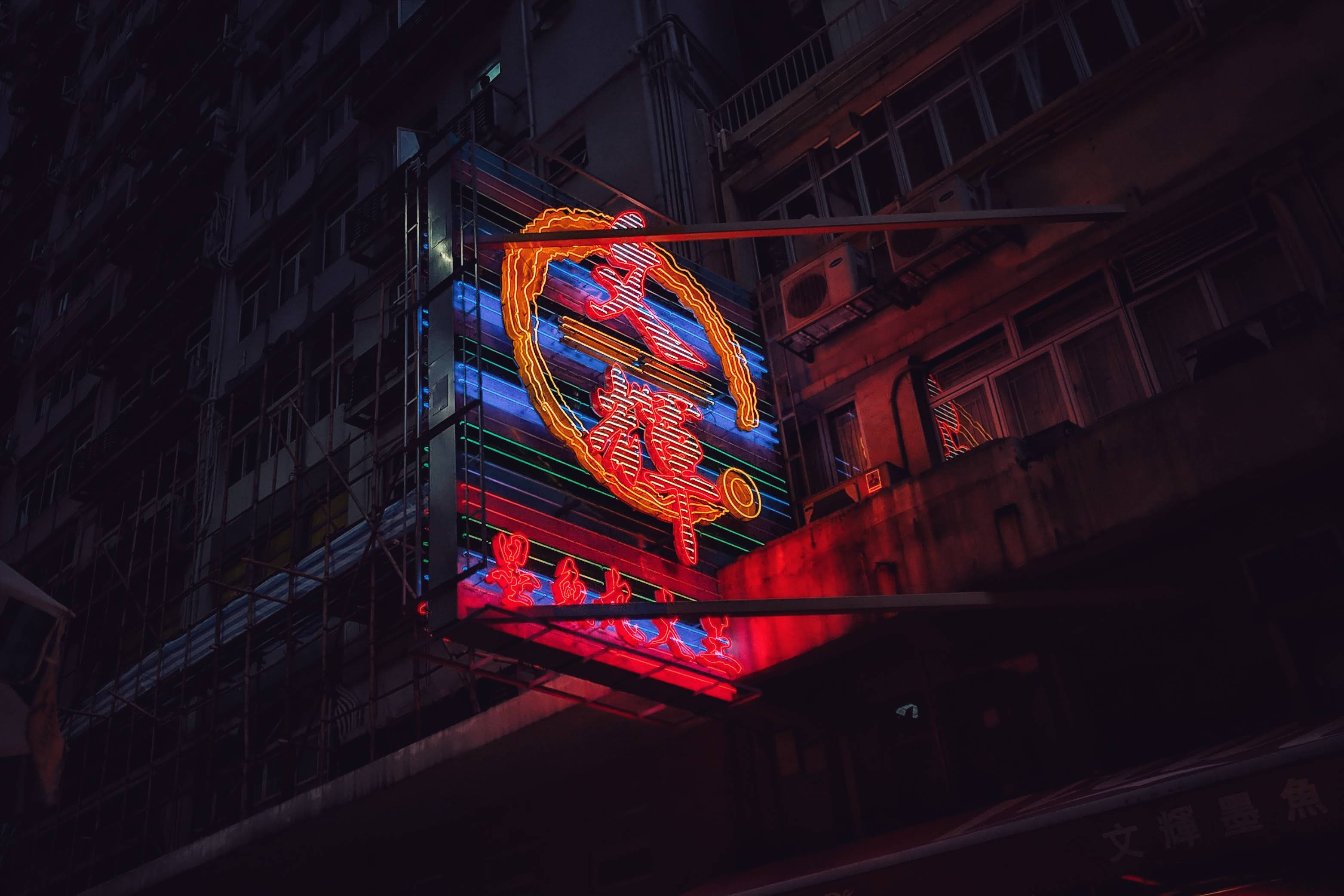 dying 桌布 壁紙 — 夜間，一個充滿活力的霓虹燈牌，帶有紅色和藍色發光字元，懸掛在黑暗的城市建築物上。