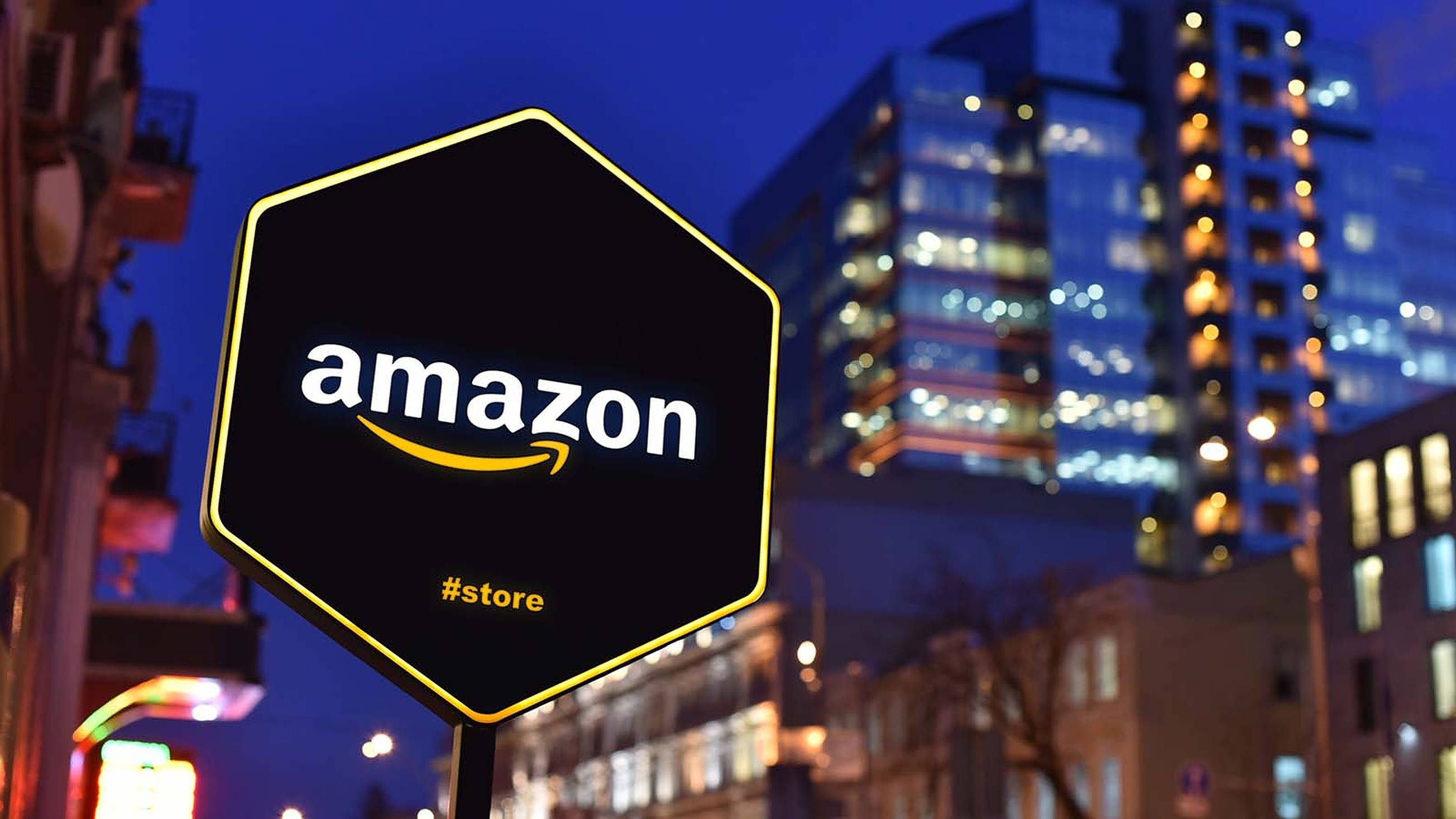 亞馬遜 桌布 壁紙 — 夜間，一個發光的六邊形Amazon商店標誌矗立在模糊的城市建築前。