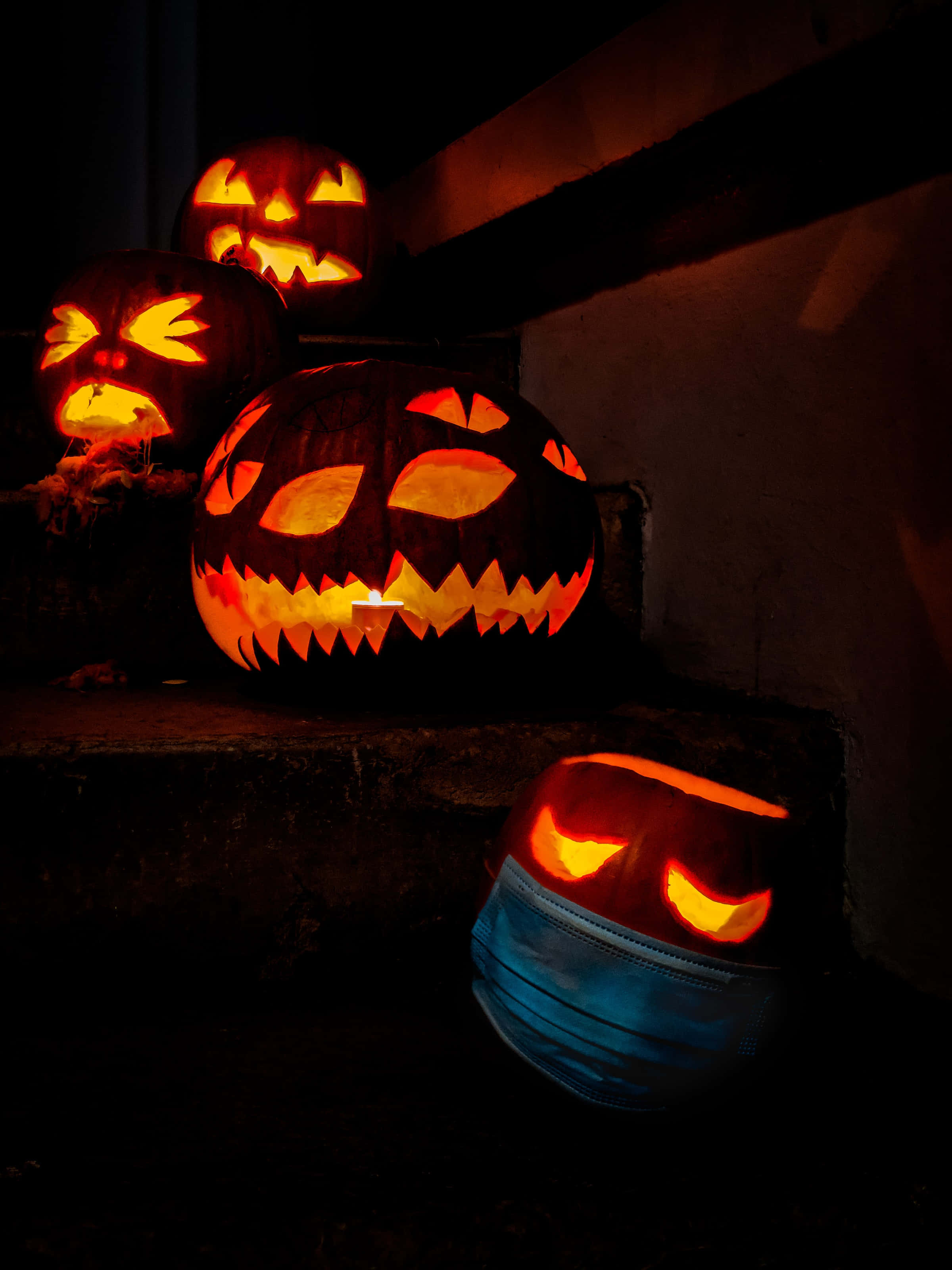 jack o'lantern 桌布 壁紙 — 夜間，發光的南瓜燈籠帶有威脅性的面孔，其中一個戴著藍色外科口罩，放在深色石階上。