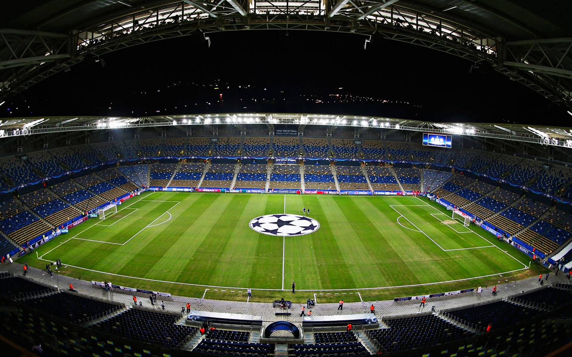 Stadium 桌布 壁紙 — 夜間一個明亮照明的足球場的廣闊鳥瞰圖，空藍色座位和綠色球場中央的UEFA Champions League標誌。