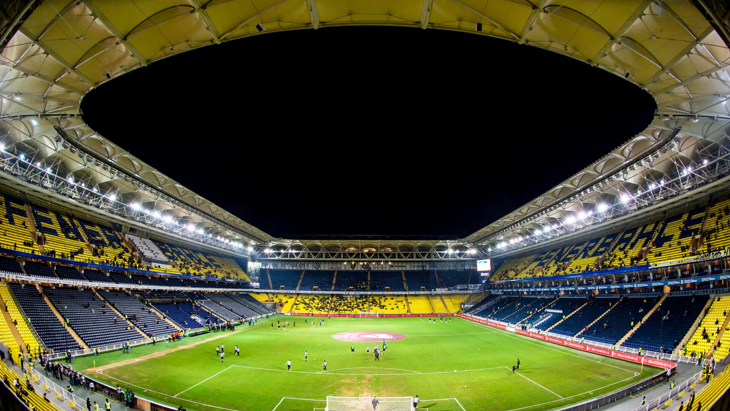 Soccer Stadium 桌布 壁紙 — 夜間一個燈火通明的足球場的廣角魚眼視圖，場上有球員，黃色和藍色座位上有球迷。