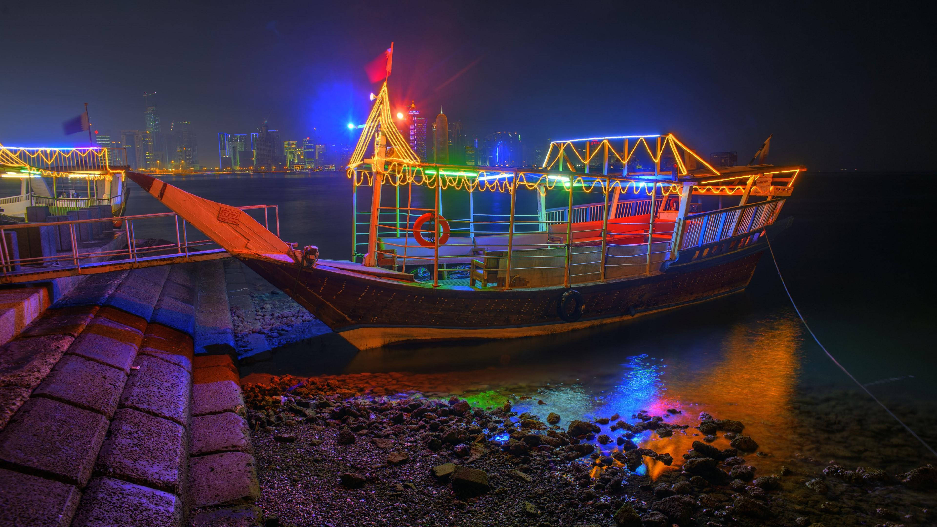 夜空 桌布 壁紙 — 夜間停泊的傳統單桅帆船，燈光璀璨，背景是閃爍的城市天際線。