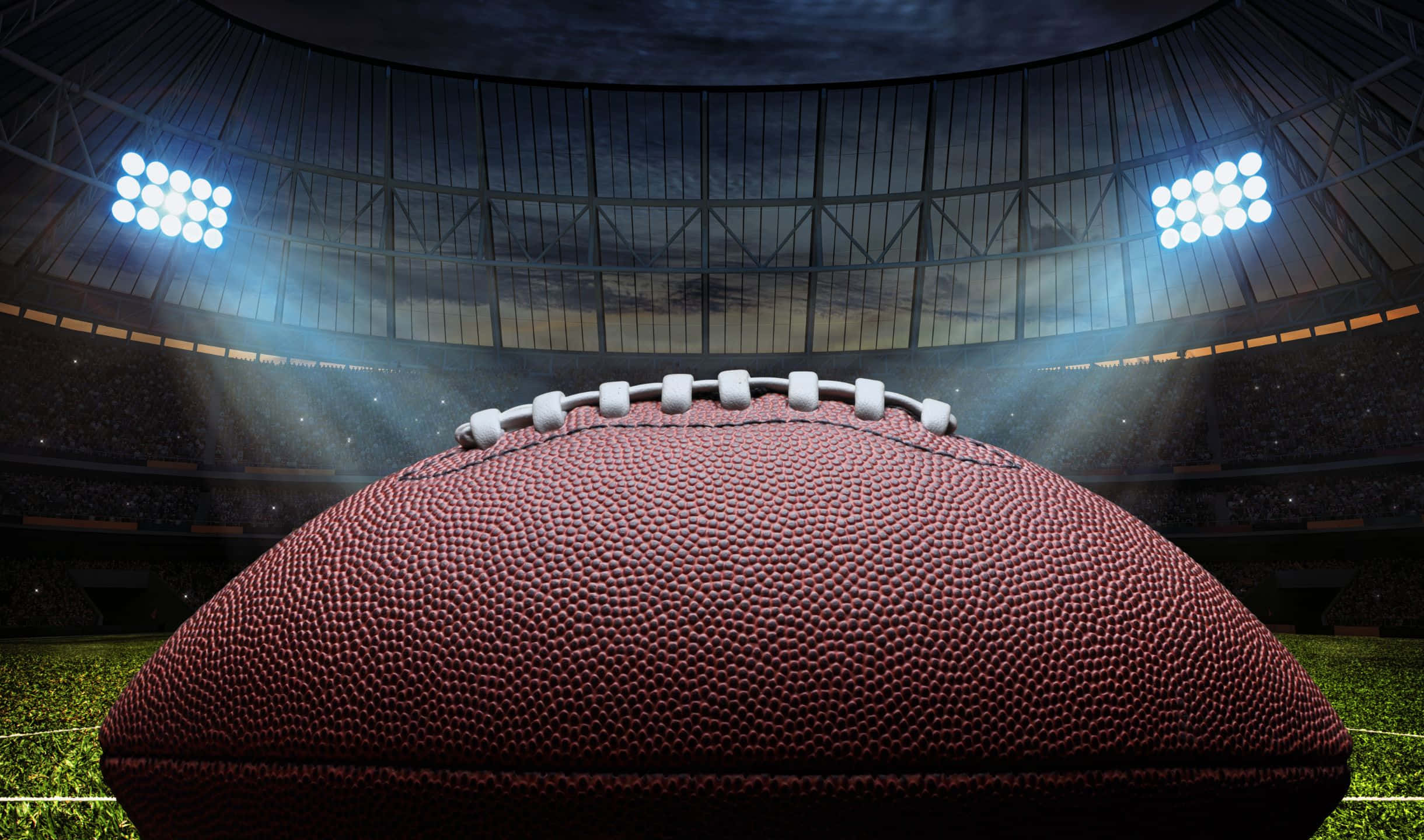 2440x1440 Football 桌布 壁紙 — 夜間在明亮球場燈光下，一個棕色皮革足球的特寫，放置在綠色草地上。