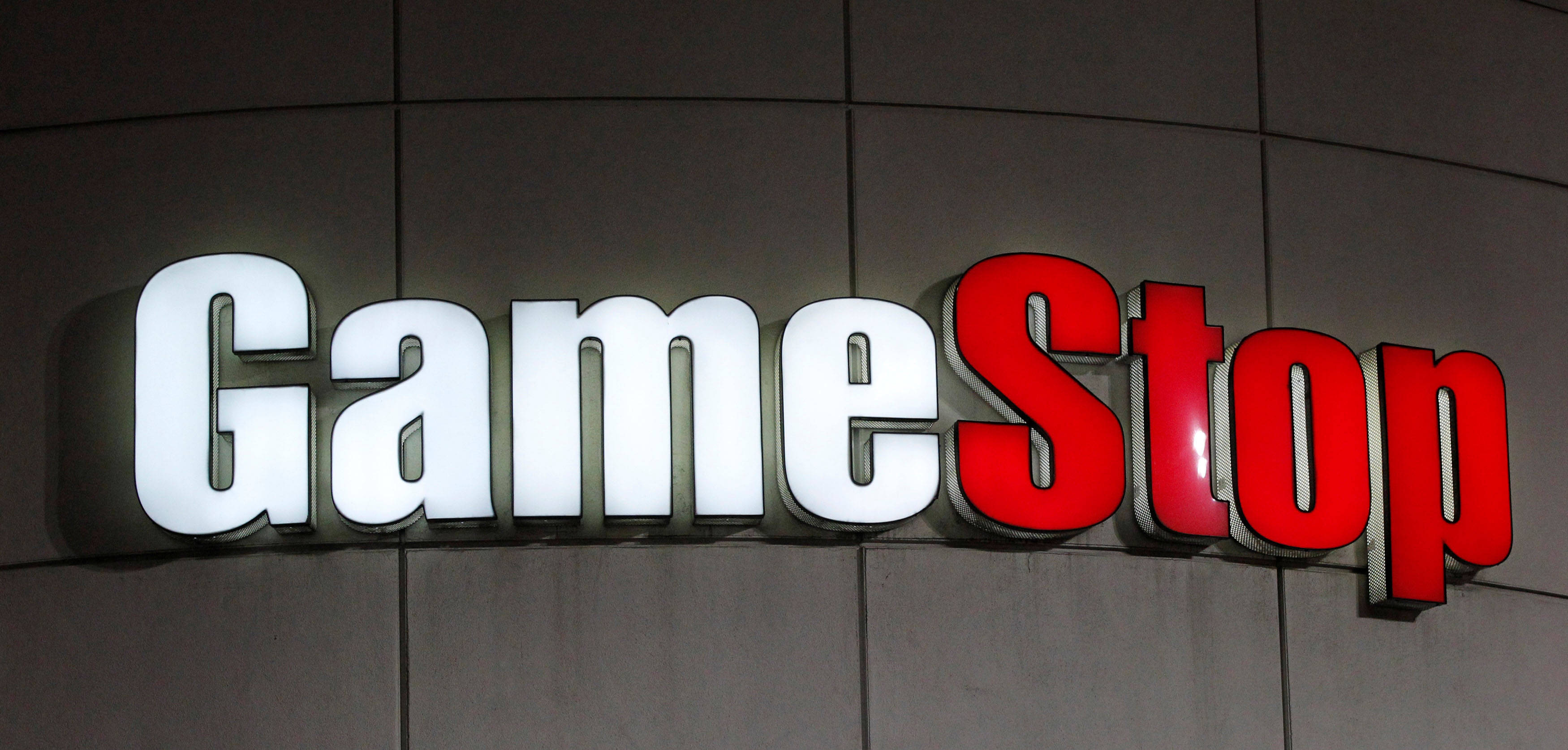 GameStop 桌布 壁紙 — 夜間安裝在深色瓷磚牆上的GameStop標誌，白色和紅色字母明亮照明。